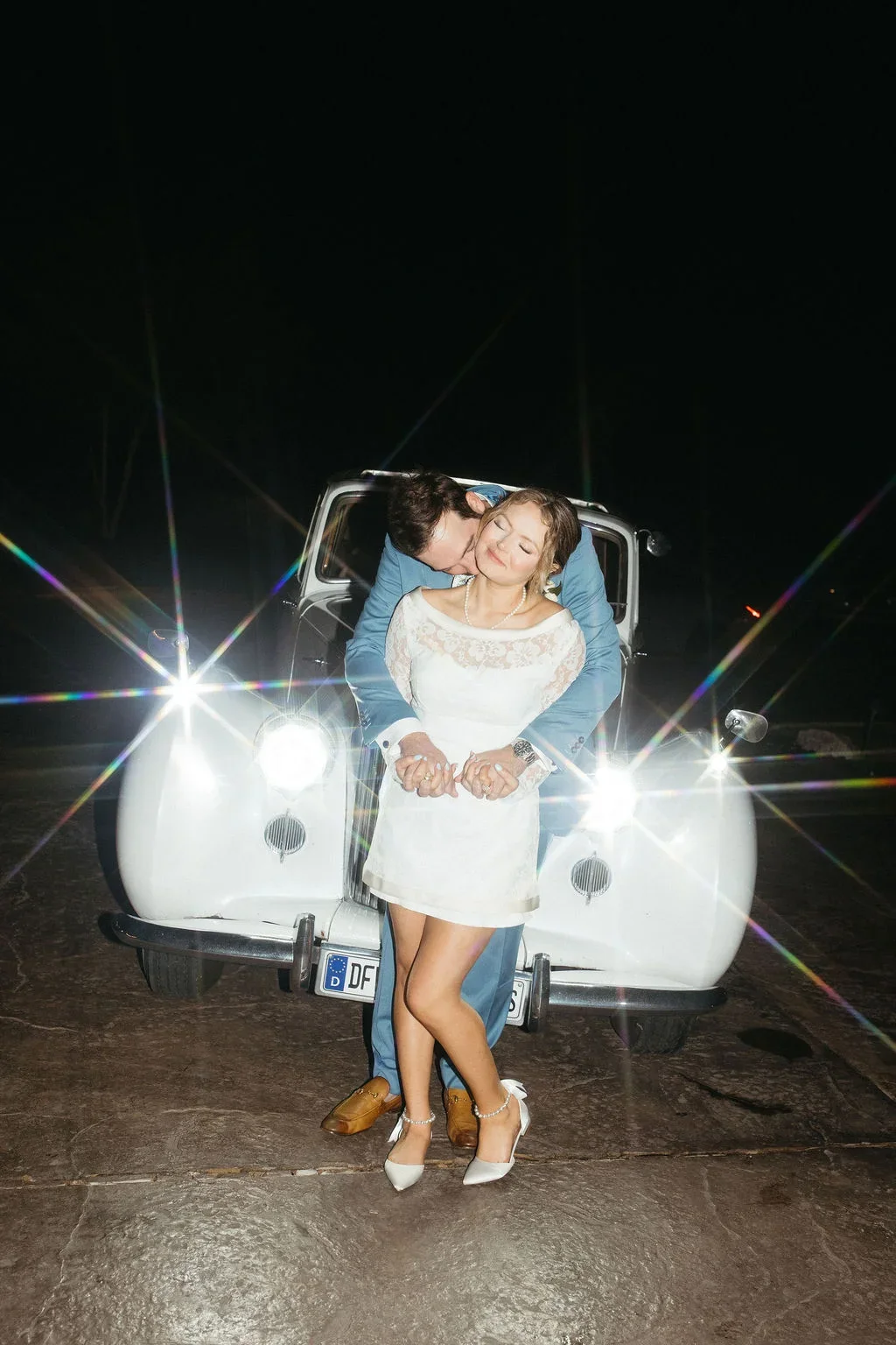 A couple dressed in wedding attire sharing an intimate moment in front of a vintage white car at night, with bright lights creating starbursts.