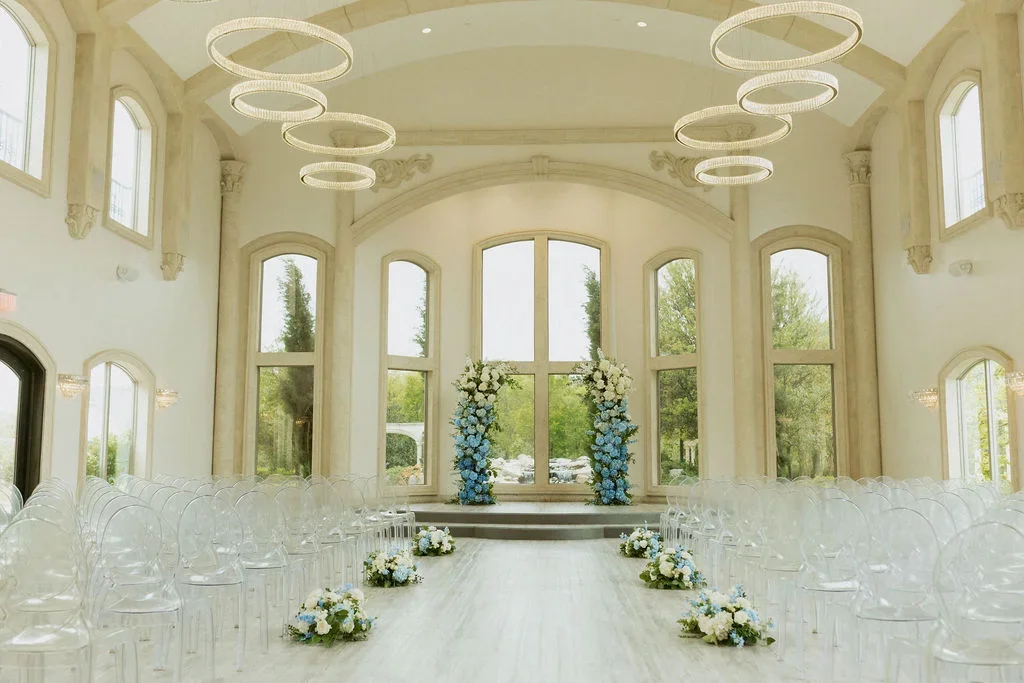 Empty wedding ceremony room with floral arrangements, high windows, and modern chandeliers.