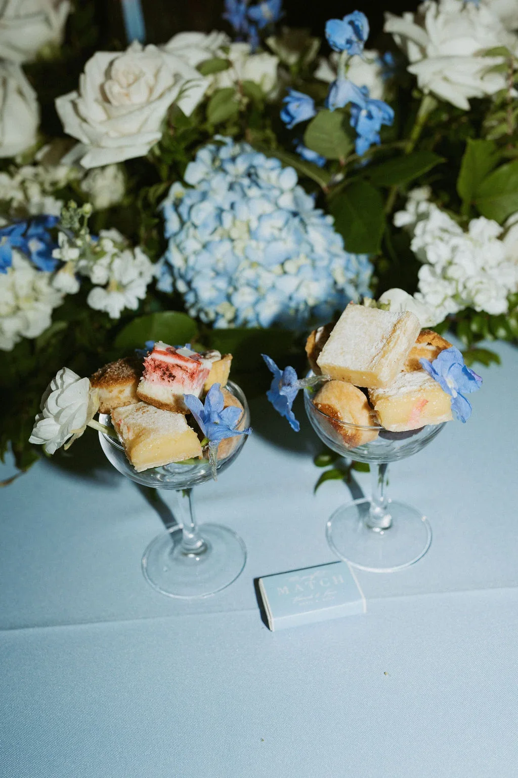 Two glass dessert cups filled with assorted petit fours decorated with blue flowers, placed on a white tablecloth in front of a floral arrangement of white and blue hydrangeas and roses.