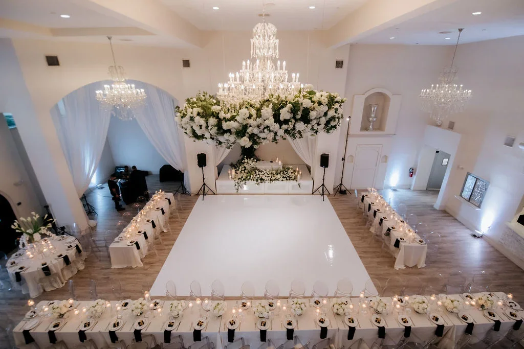 Elegant wedding reception hall with white decor, floral arrangements, chandeliers, and a dance floor.