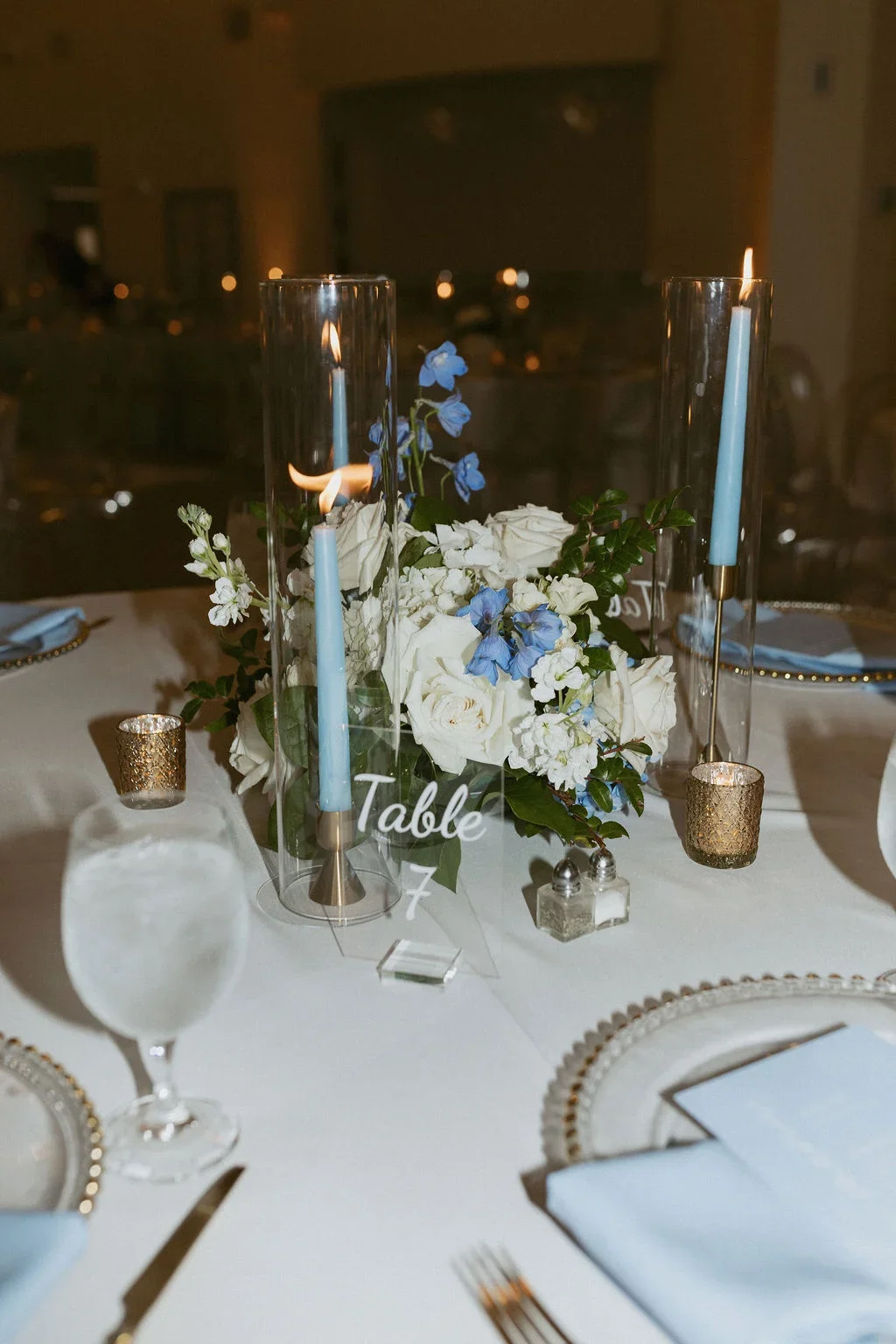 Elegant table centerpiece with white roses, blue flowers, and green foliage in tall glass vases, with blue taper candles and gold votive candles, set for a formal event.