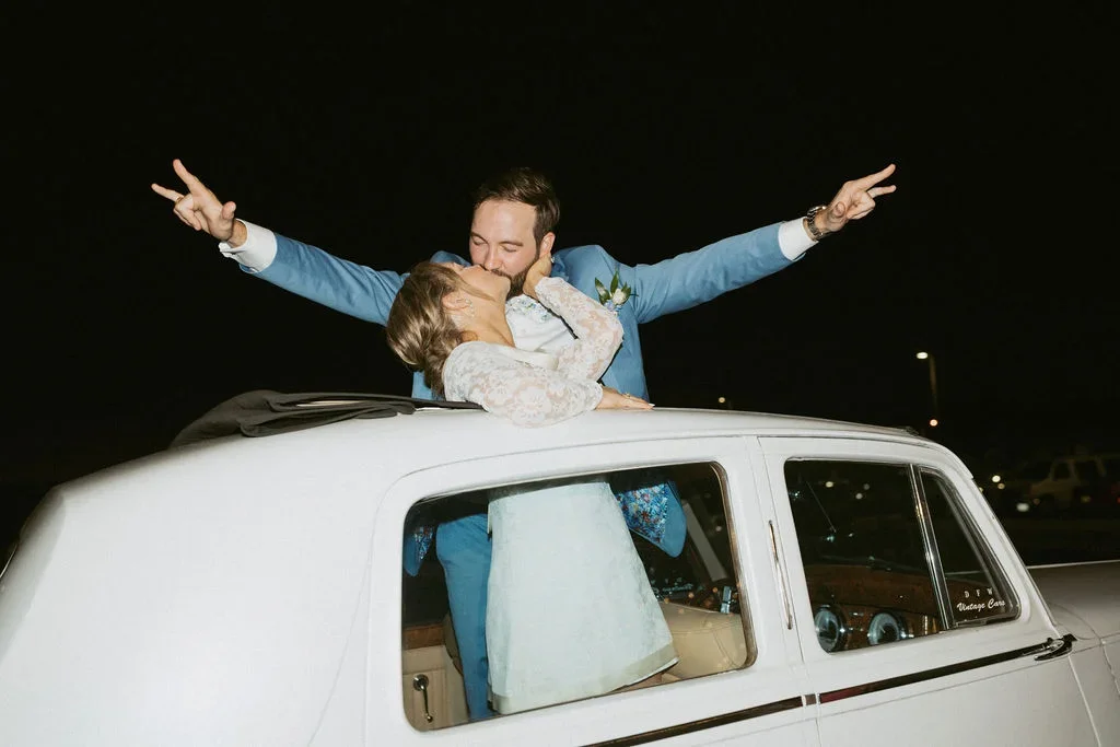 A groom and bride sharing a kiss at night in a vintage car, with the groom making a peace sign with both hands, celebrating their wedding.