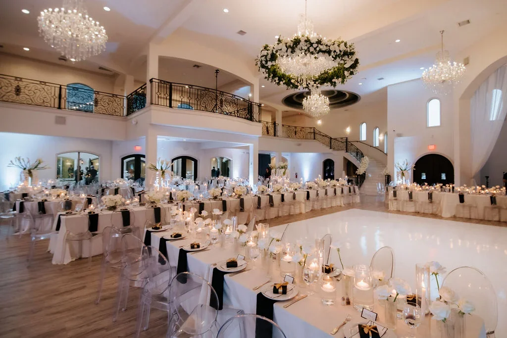 Elegant wedding reception hall with round tables, white tablecloths with black accents, flower centerpieces, candles, and clear ghost chairs. Chandeliers hang from the ceiling, and there is a dance floor in the center. The room features a second-floo