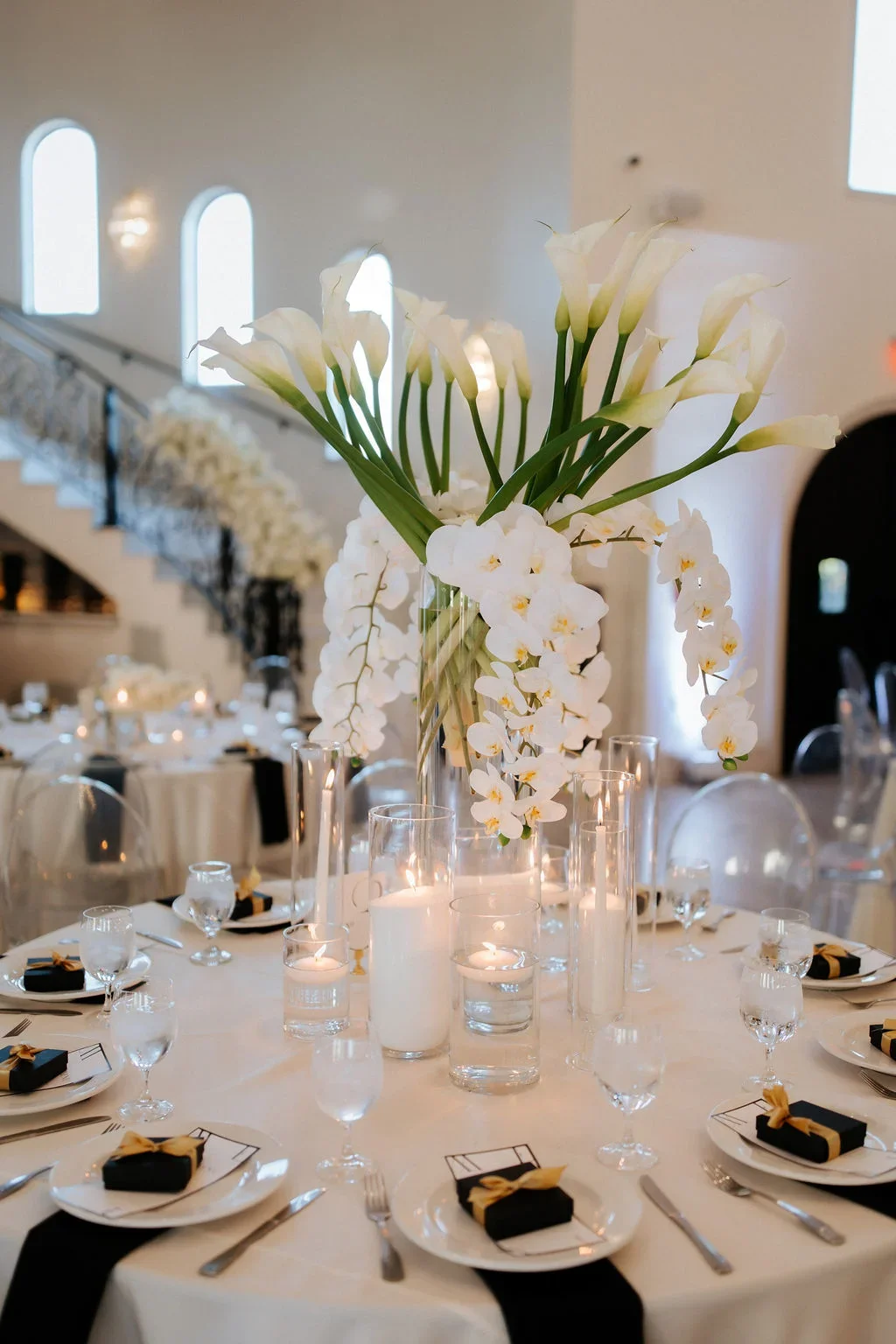 Elegant banquet table centerpiece with white orchids and calla lilies in a tall glass vase, surrounded by candles in glass holders, set for a formal event.