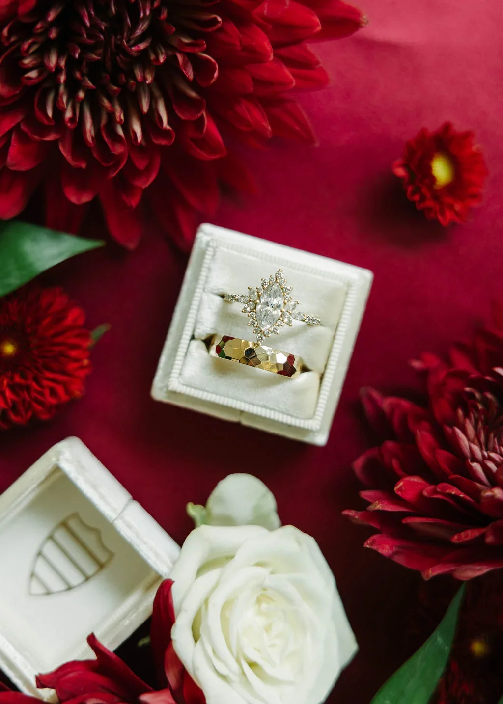 A pair of rings displayed in a white jewelry box, surrounded by red and white flowers on a dark red surface.