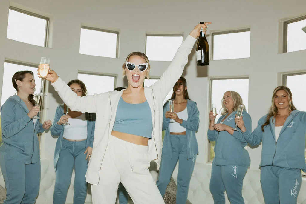 Group of women in matching blue pajamas celebrating at a party, with one woman in front wearing heart-shaped sunglasses, holding a champagne bottle and glasses, in a bright living room with large windows.