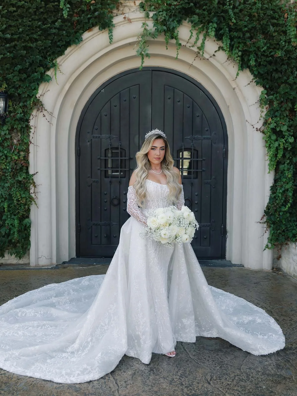 A bride in a white wedding dress holding a bouquet of white roses, standing in front of a black arched gate with greenery.