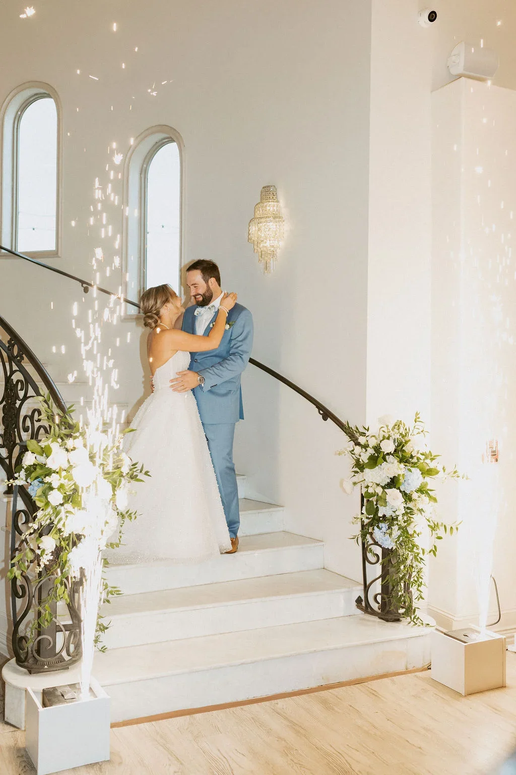 A bride and groom are embracing and smiling on a staircase during their wedding celebration, with sparklers and floral arrangements nearby.