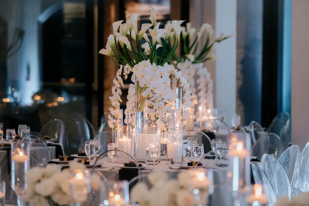 Elegant banquet table setup with tall floral centerpieces of white flowers and greenery, candles, glassware, and transparent chairs.