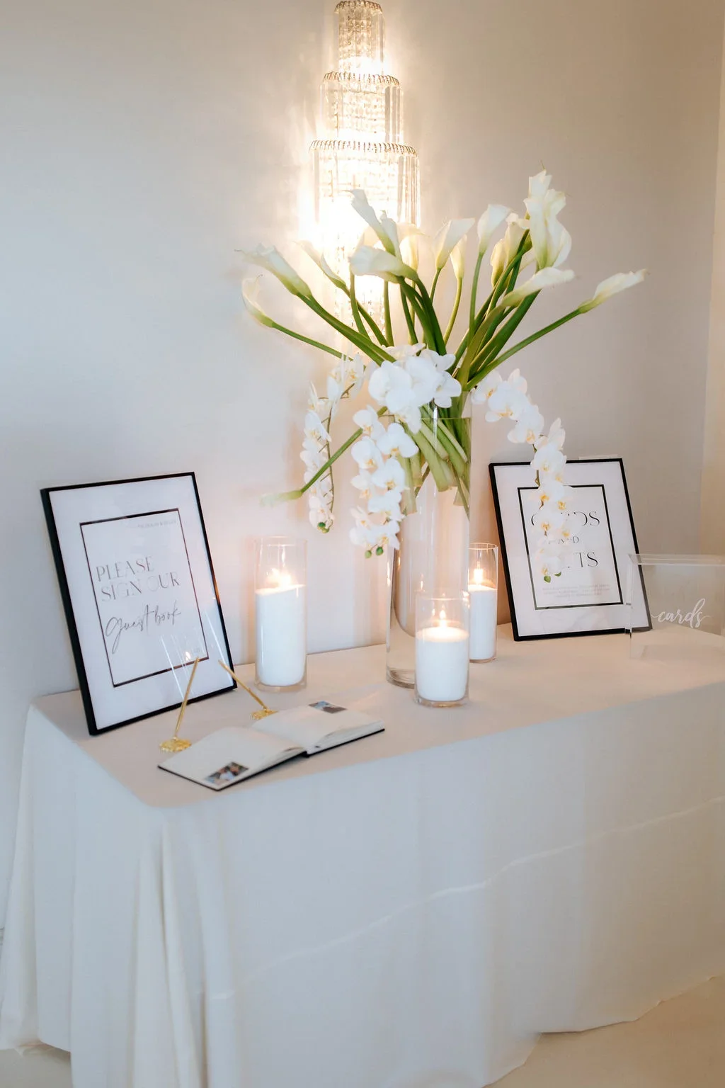 A table decorated with white flowers in a tall vase, surrounded by candles, sign-in sheets, and framed signs at a wedding or event.
