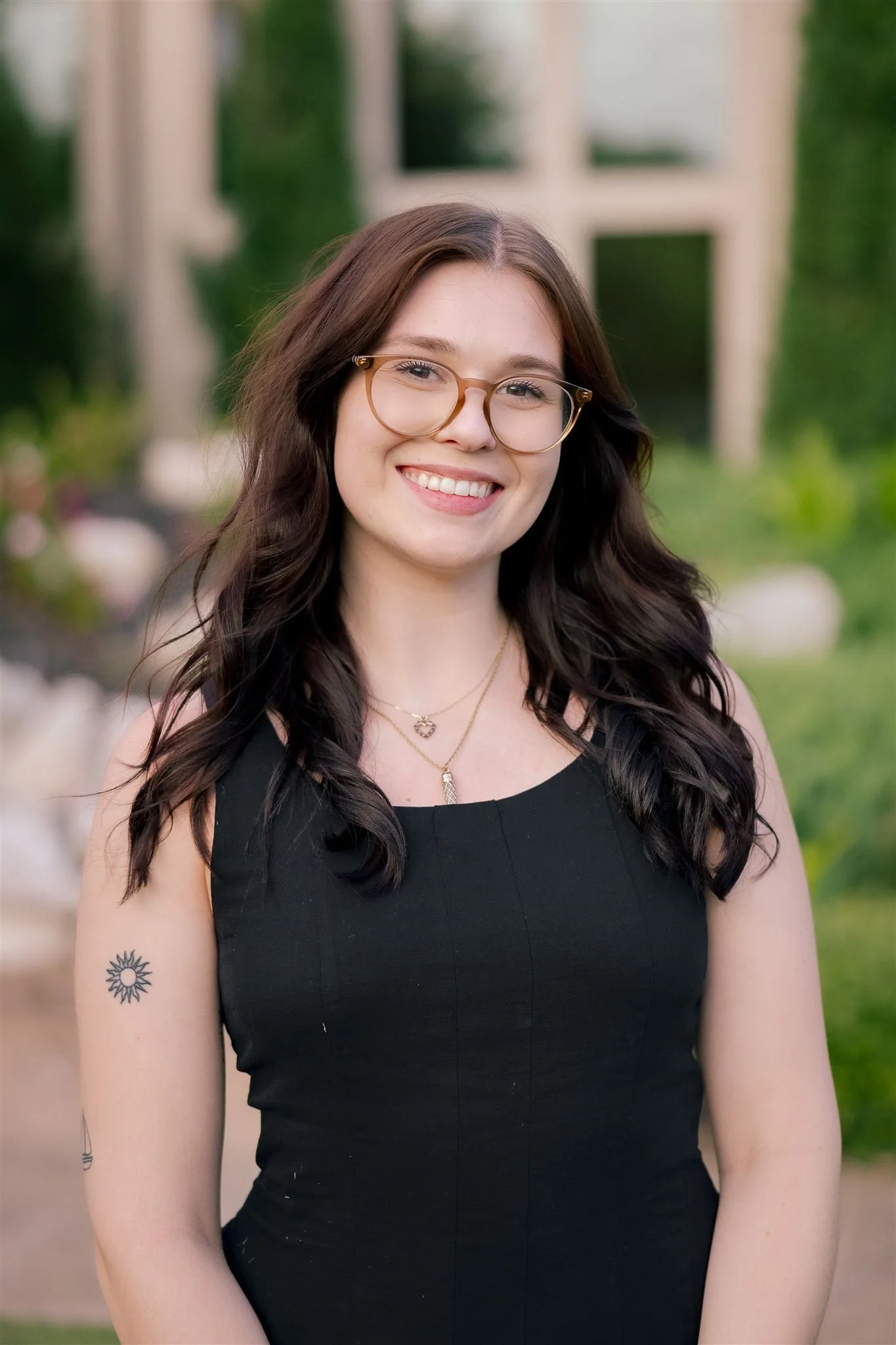 Smiling young woman with long dark hair and glasses, wearing a black sleeveless top and layered necklaces, standing outdoors with greenery and a structure in the background.