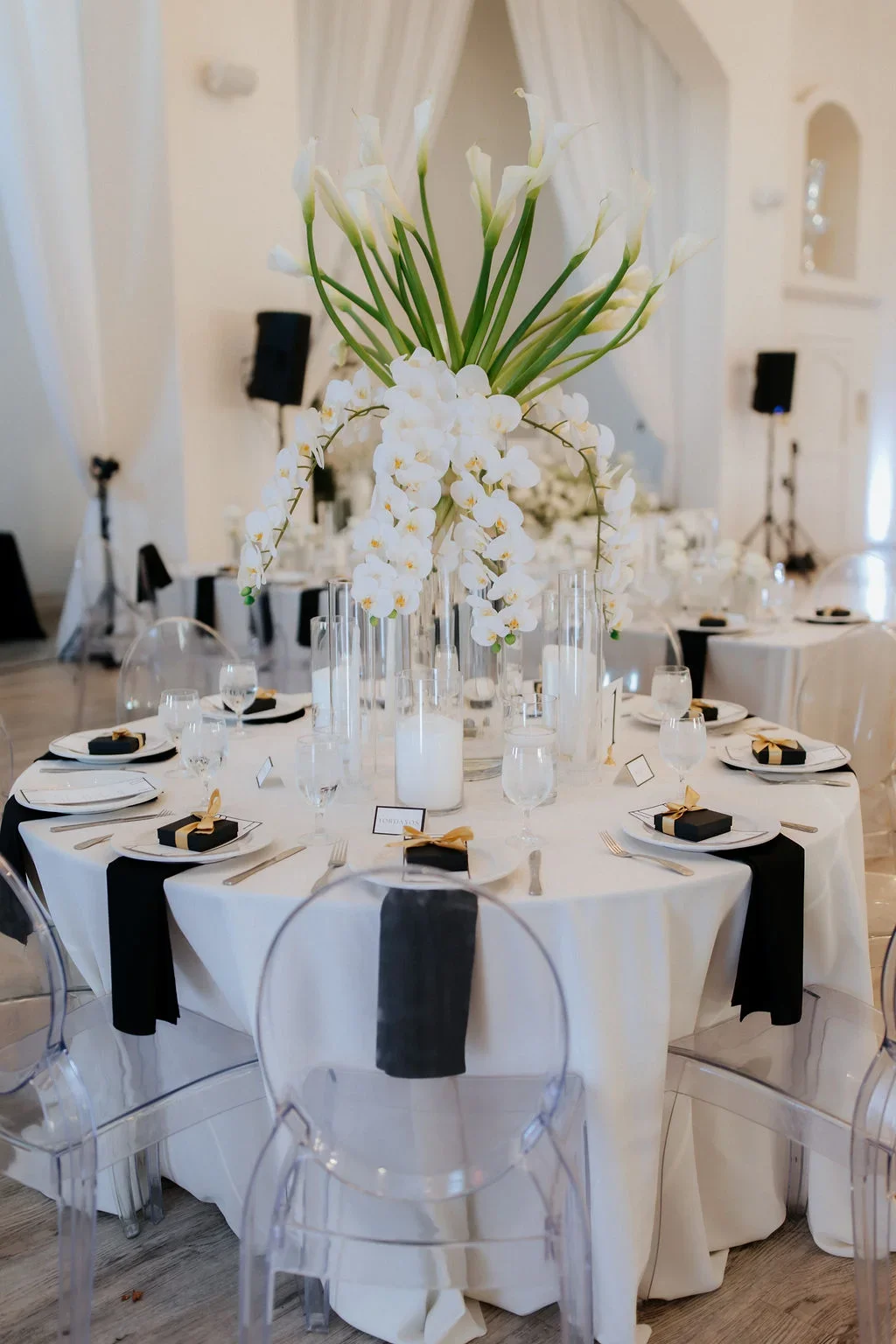Elegant round table set for a formal event with a tall floral centerpiece of white orchids and calla lilies, surrounded by glassware, black and white napkins, and small black gift boxes with gold ribbons, in a decorated banquet hall.