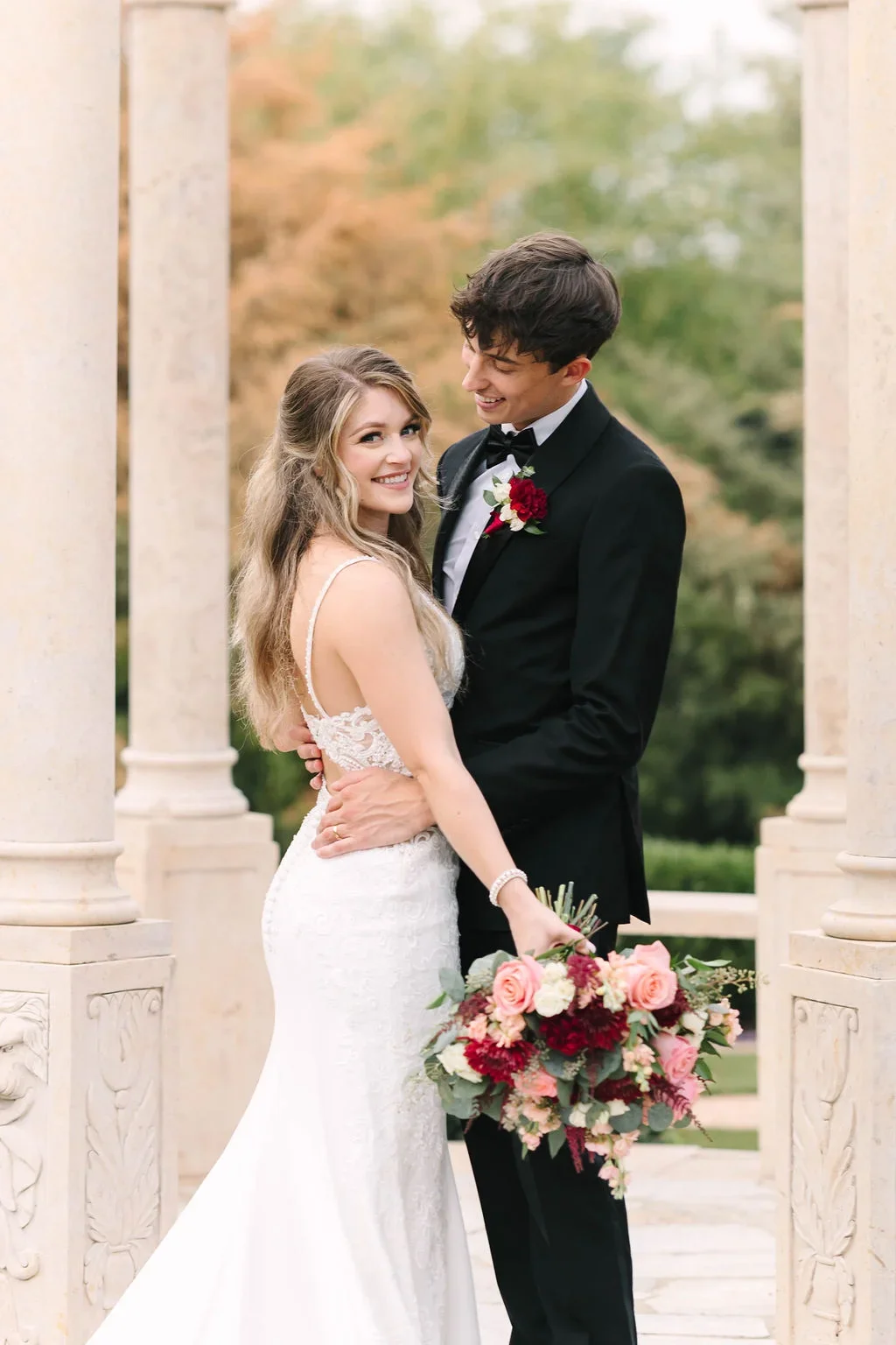 A bride and groom standing close, smiling, with the bride holding a bouquet of pink and red flowers, in an outdoor setting with trees and stone columns.