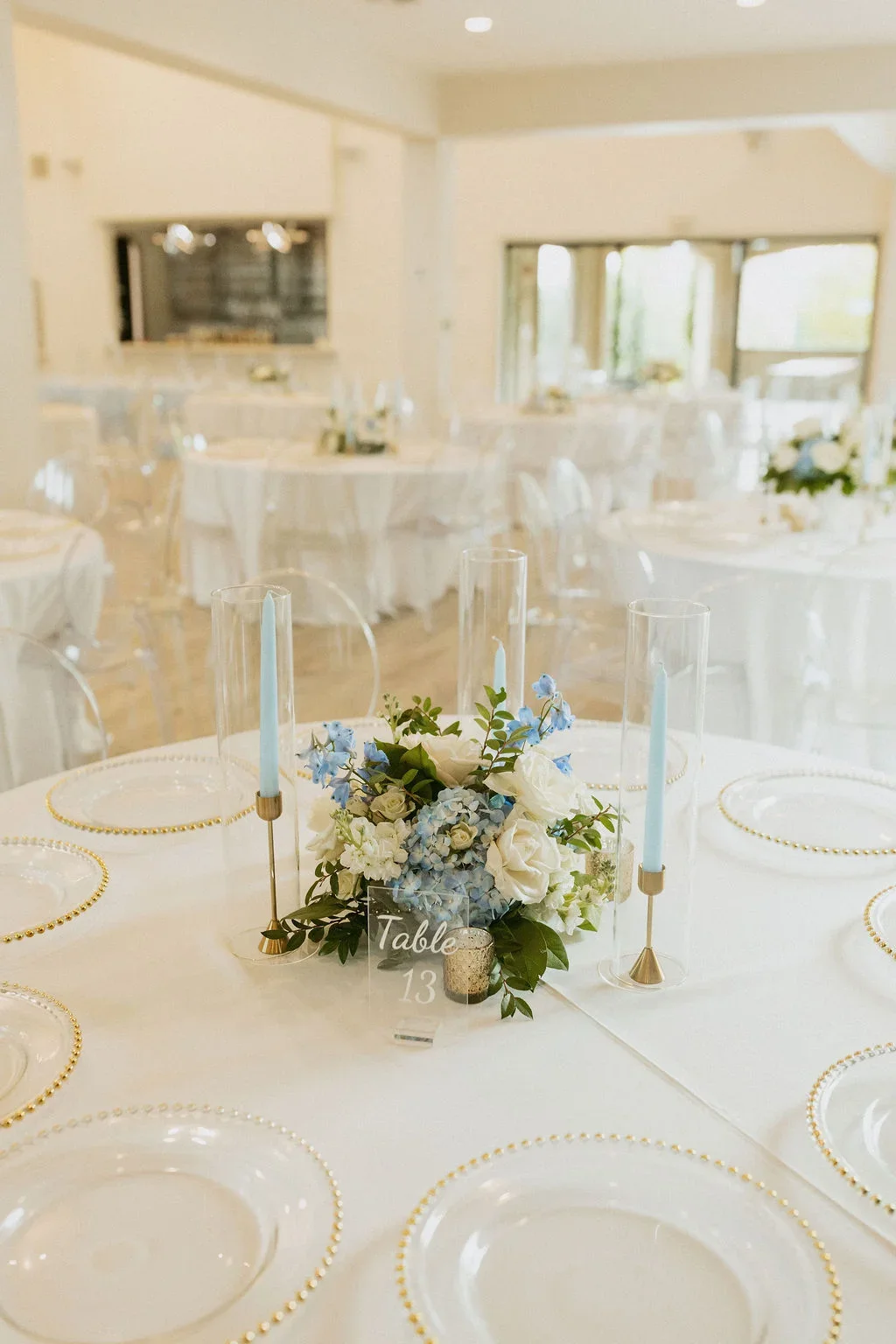 Elegant wedding reception table with white plates that have gold beads on the rim, a floral centerpiece with white and blue flowers, light blue taper candles in glass holders, and a table number sign that reads 'Table 13', in a bright room with large
