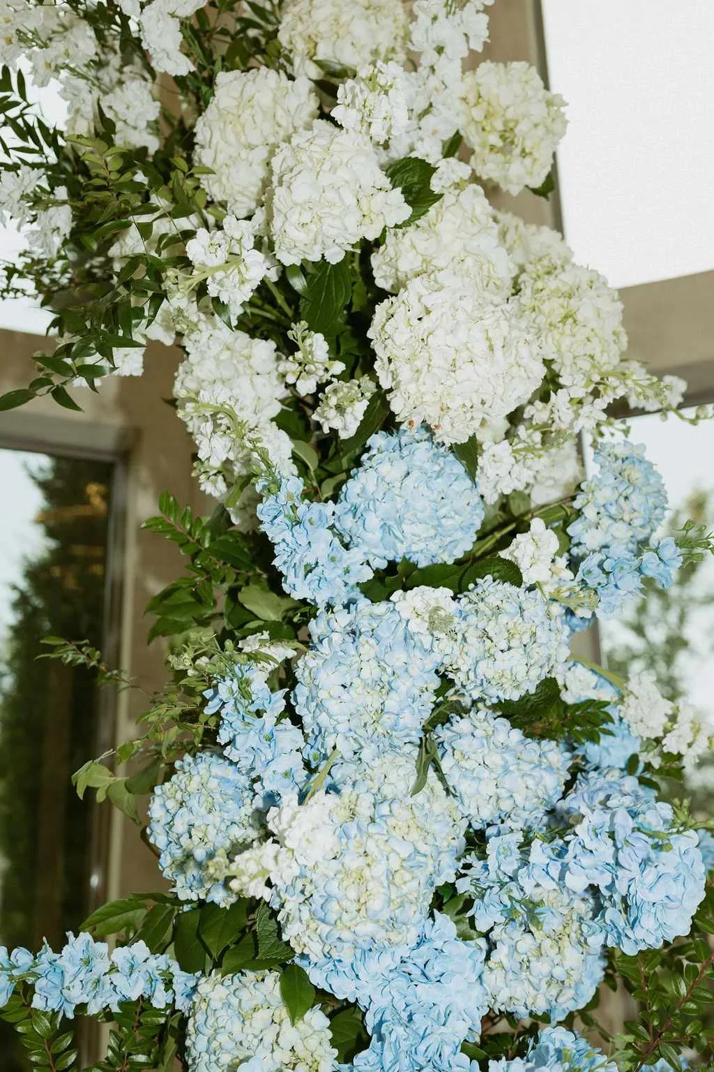 A large bouquet of white and light blue hydrangea flowers with green leaves, hanging outdoors near a building.