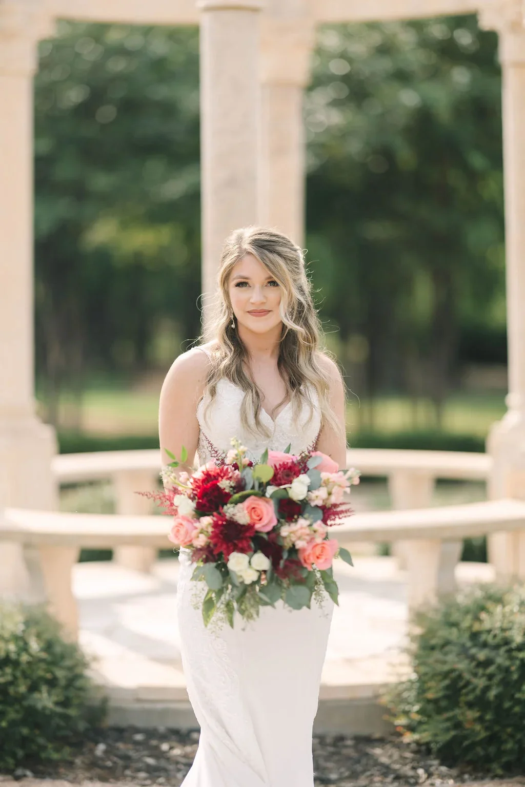 Bride in a white wedding dress holding a large bouquet of pink, red, and white flowers outdoors.