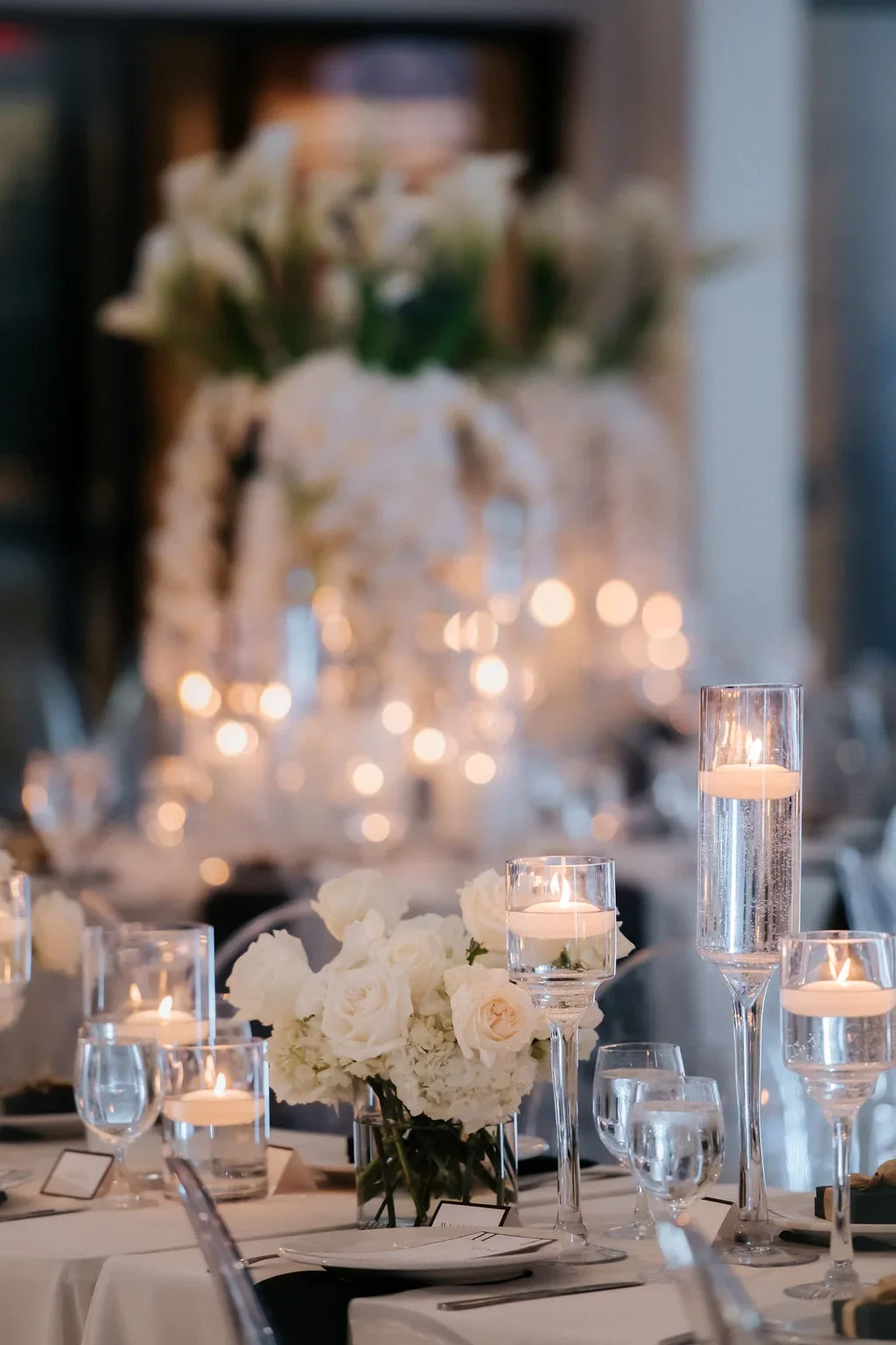 Elegant table setting with white roses and floating candles in glass holders, decorated for a formal event or wedding reception.