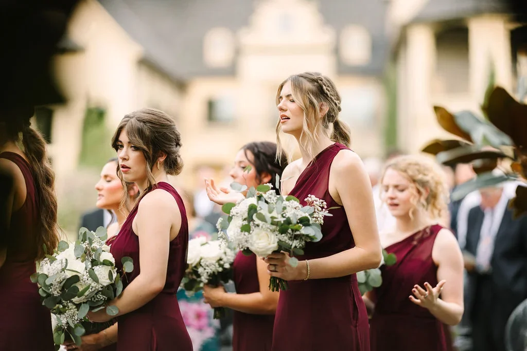 A group of women in burgundy dresses holding bouquets of white flowers during a wedding ceremony outdoors.