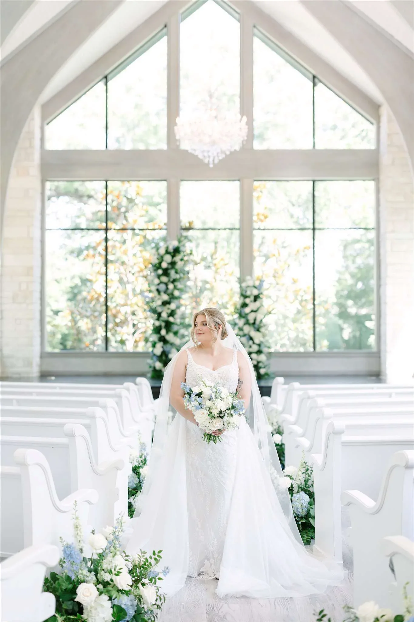 A bride in a white wedding gown holding a bouquet in a decorated wedding venue with rows of white chairs and floral arrangements.