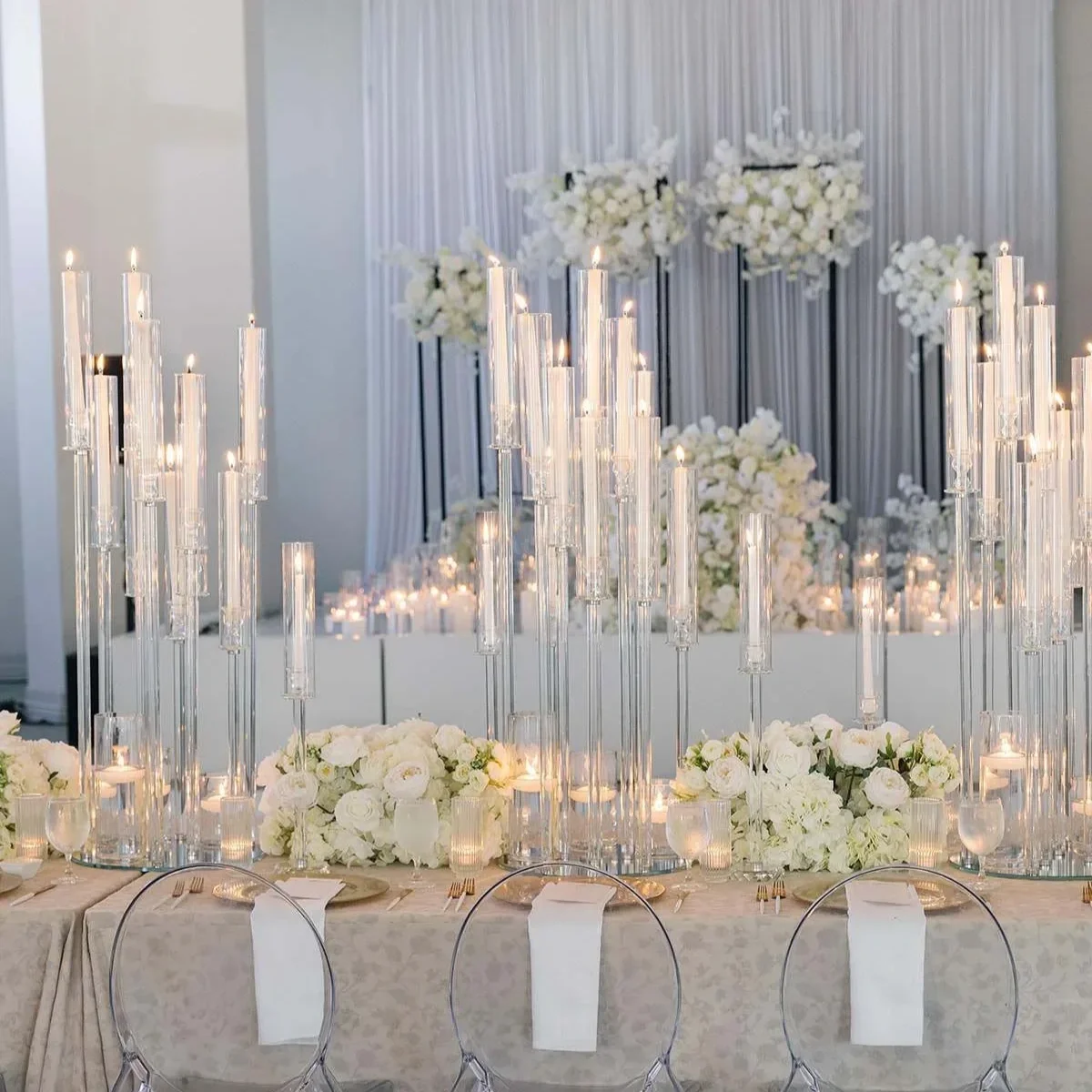 Elegant wedding reception table decorated with tall glass candle holders, white floral arrangements, and white candles, with a matching floral backdrop.