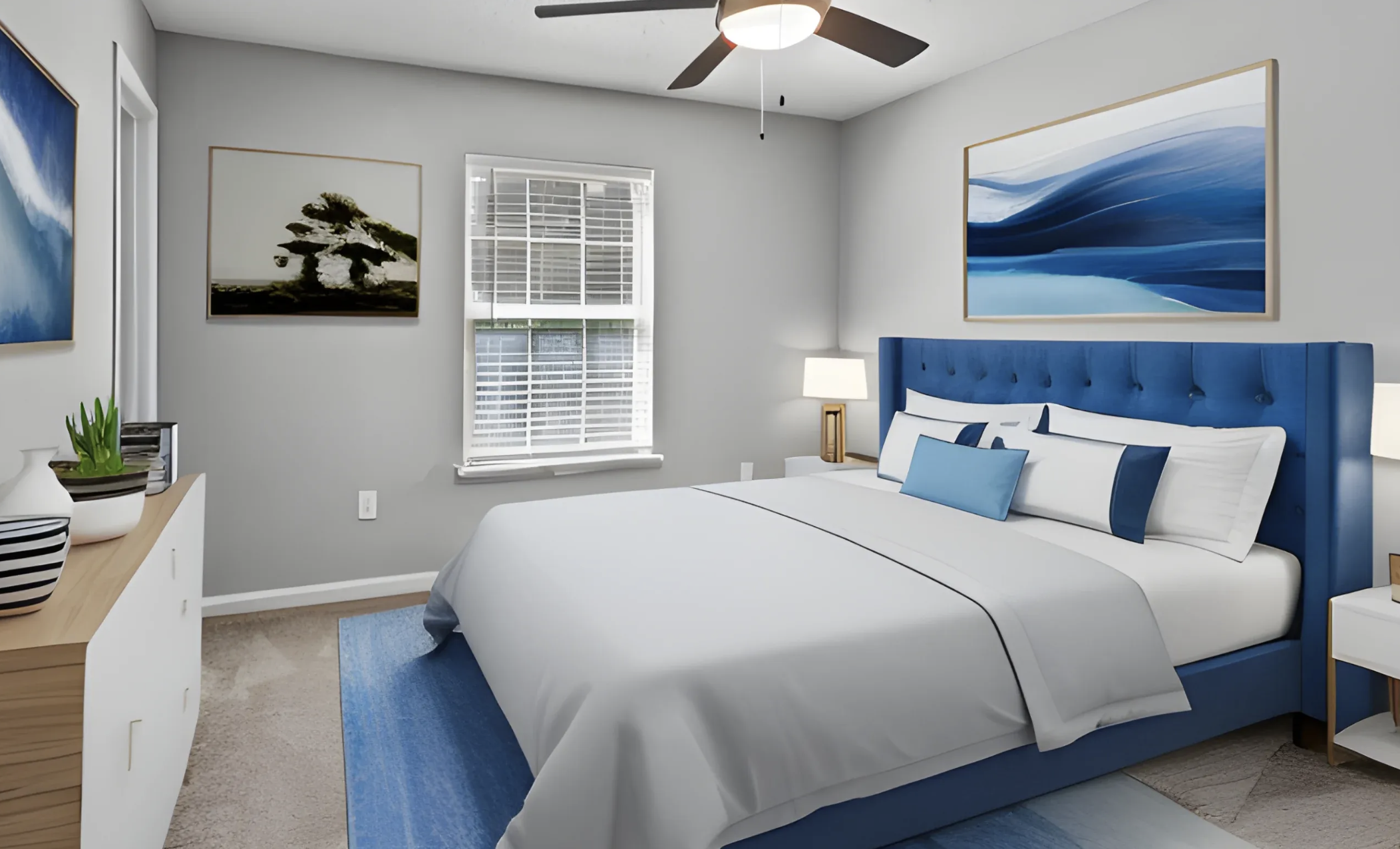 A bedroom with a blue upholstered bed, white bedding, and matching blue and white pillows, with artwork and a ceiling fan.