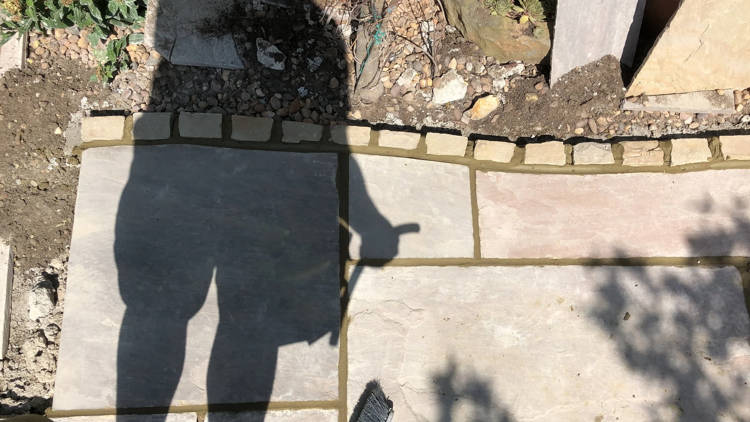 Photograph of a partially completed stone pathway with large light-colored slabs and small mortar-filled gaps, surrounded by earth, gravel, and scattered rocks, with shadows cast over the surface.