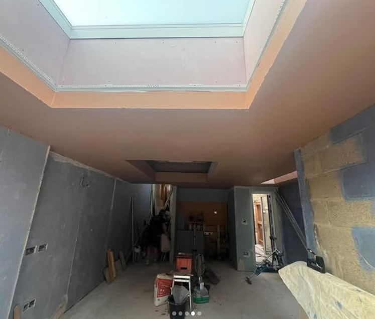 Interior of a building under construction or renovation, showing a room with unfinished walls, construction materials, and an opening in the ceiling for a skylight.
