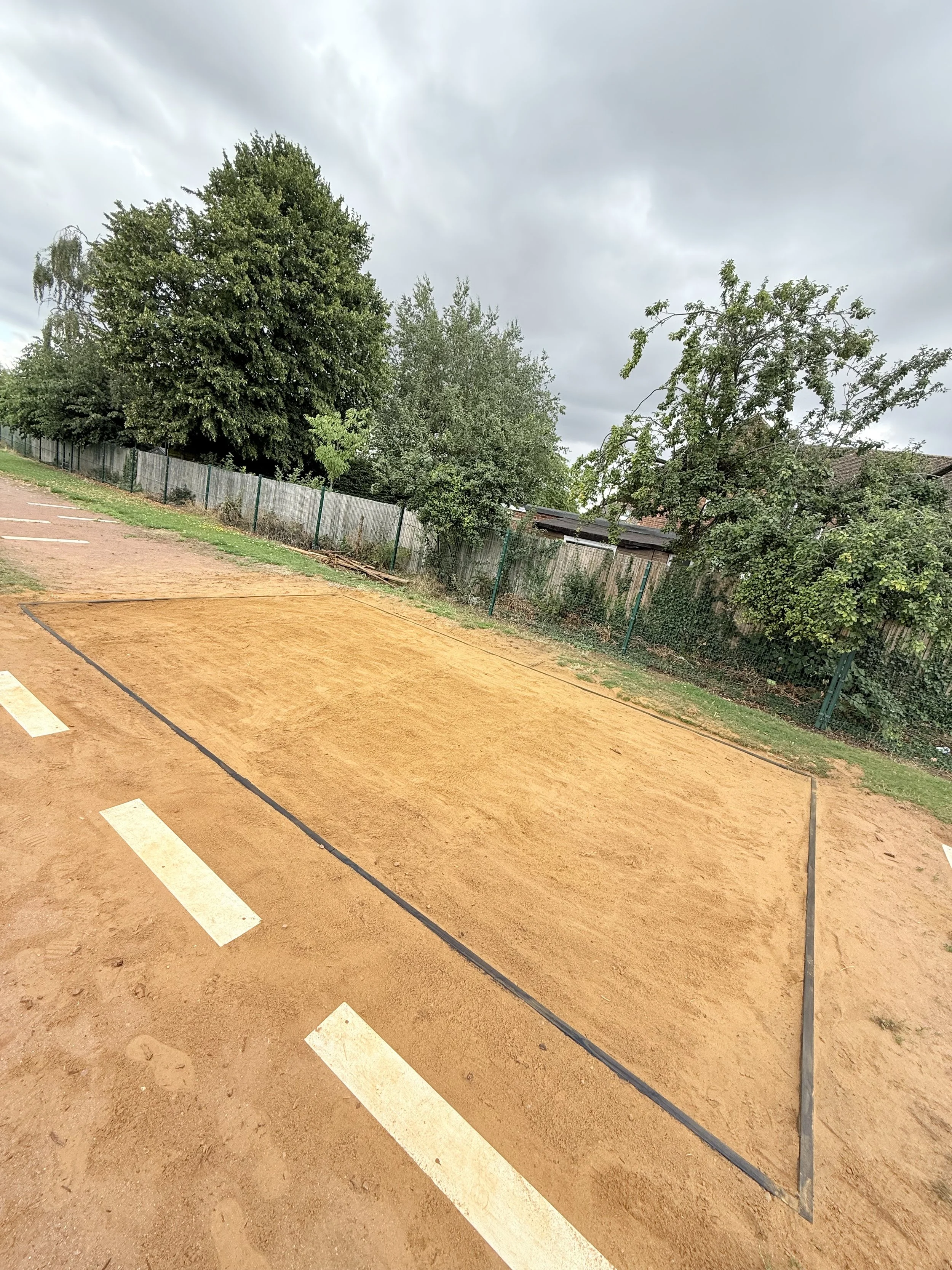 Empty sand volleyball court with boundary lines and parking spaces on the side, surrounded by a wooden fence and trees under cloudy sky.