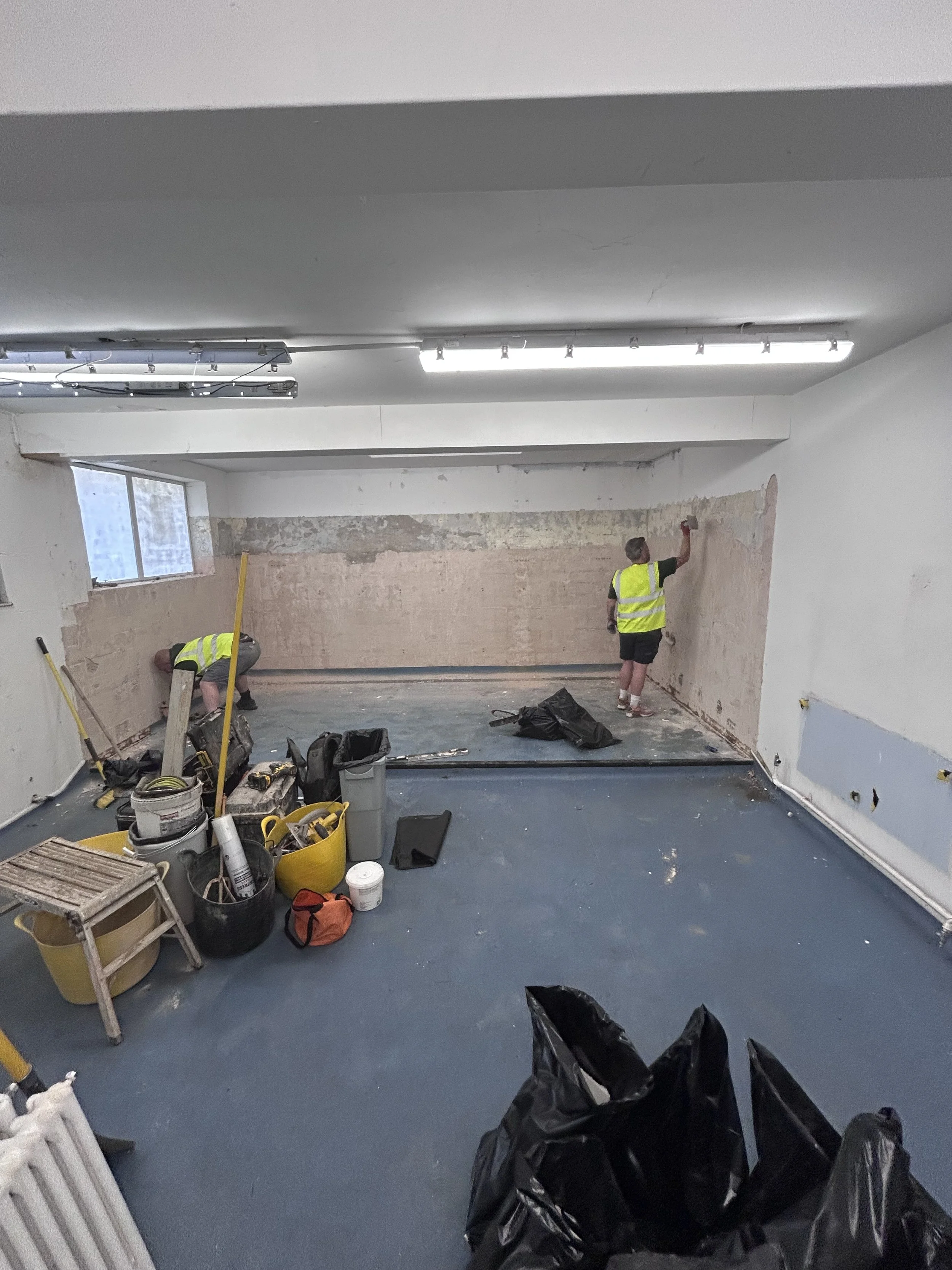 Two workers in yellow safety vests and casual clothing are renovating a room. One is sanding or scraping a wall, while the other is painting or applying a substance to the wall. The room has a window, overhead fluorescent lights, and various tools an
