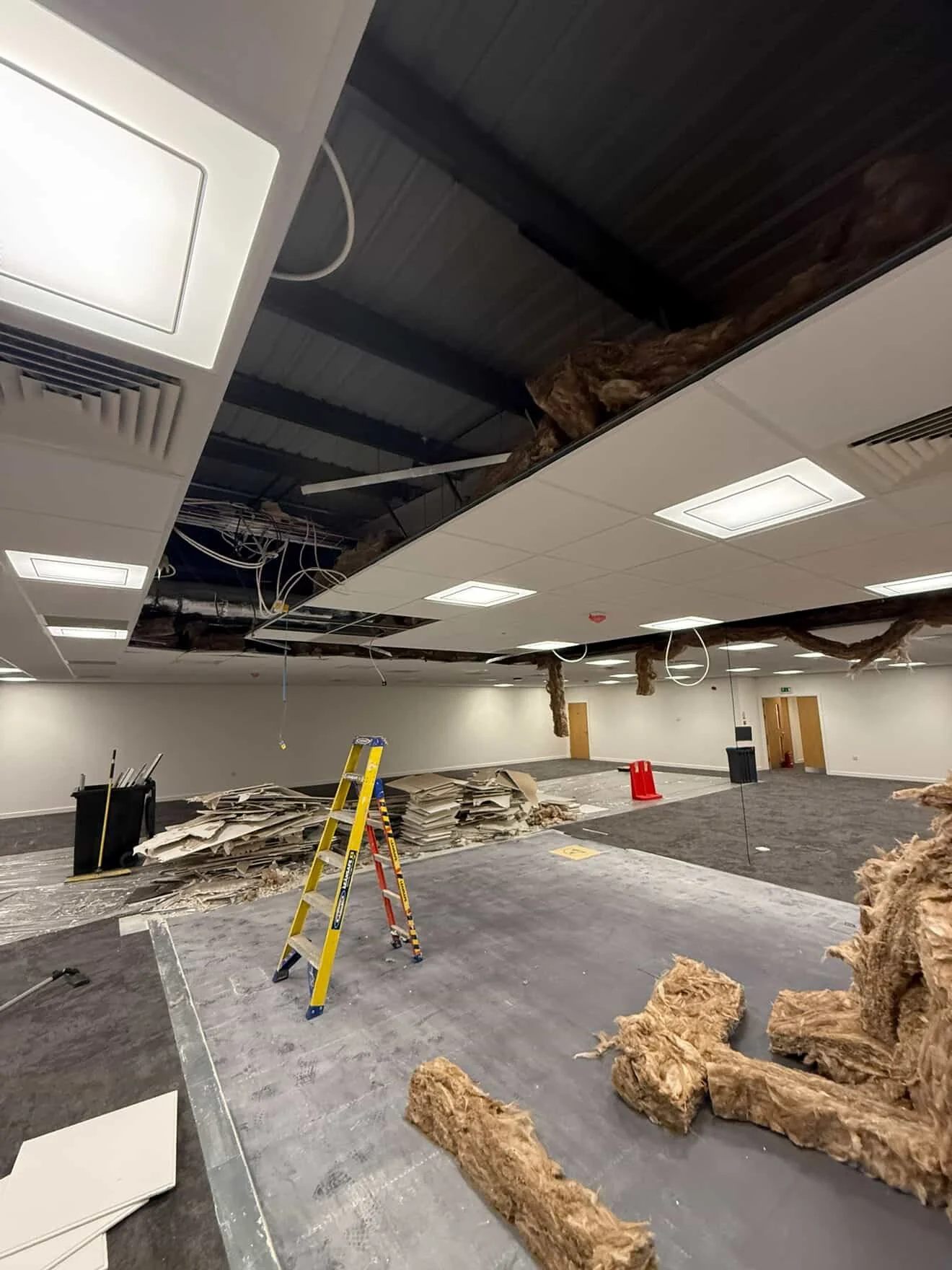Interior of a room under renovation with partially removed ceiling panels, insulation debris on the floor, a yellow ladder, construction barrels, and exposed wiring.