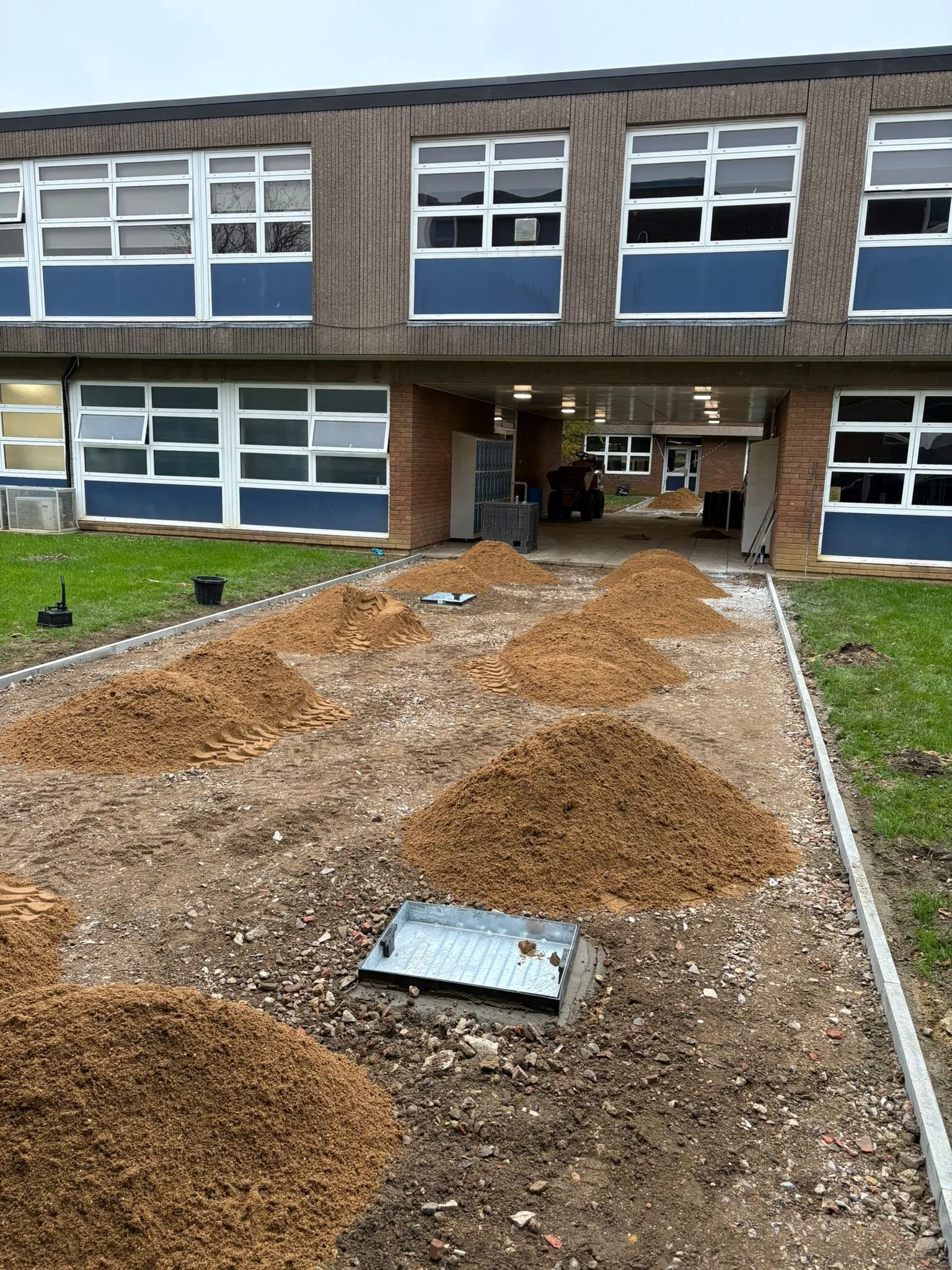 Construction site with piles of dirt and sand, gravel ground, and a metal tray in front of a building with multiple windows and an underpass.