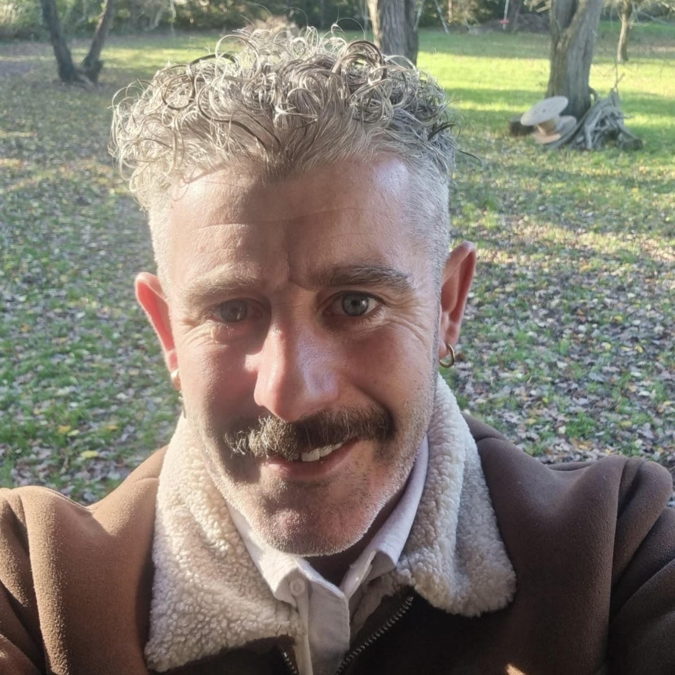 A smiling man with curly gray hair, blue eyes, a beard, and a small earring in his left ear, taking a selfie outdoors in a park with trees and fallen leaves.
