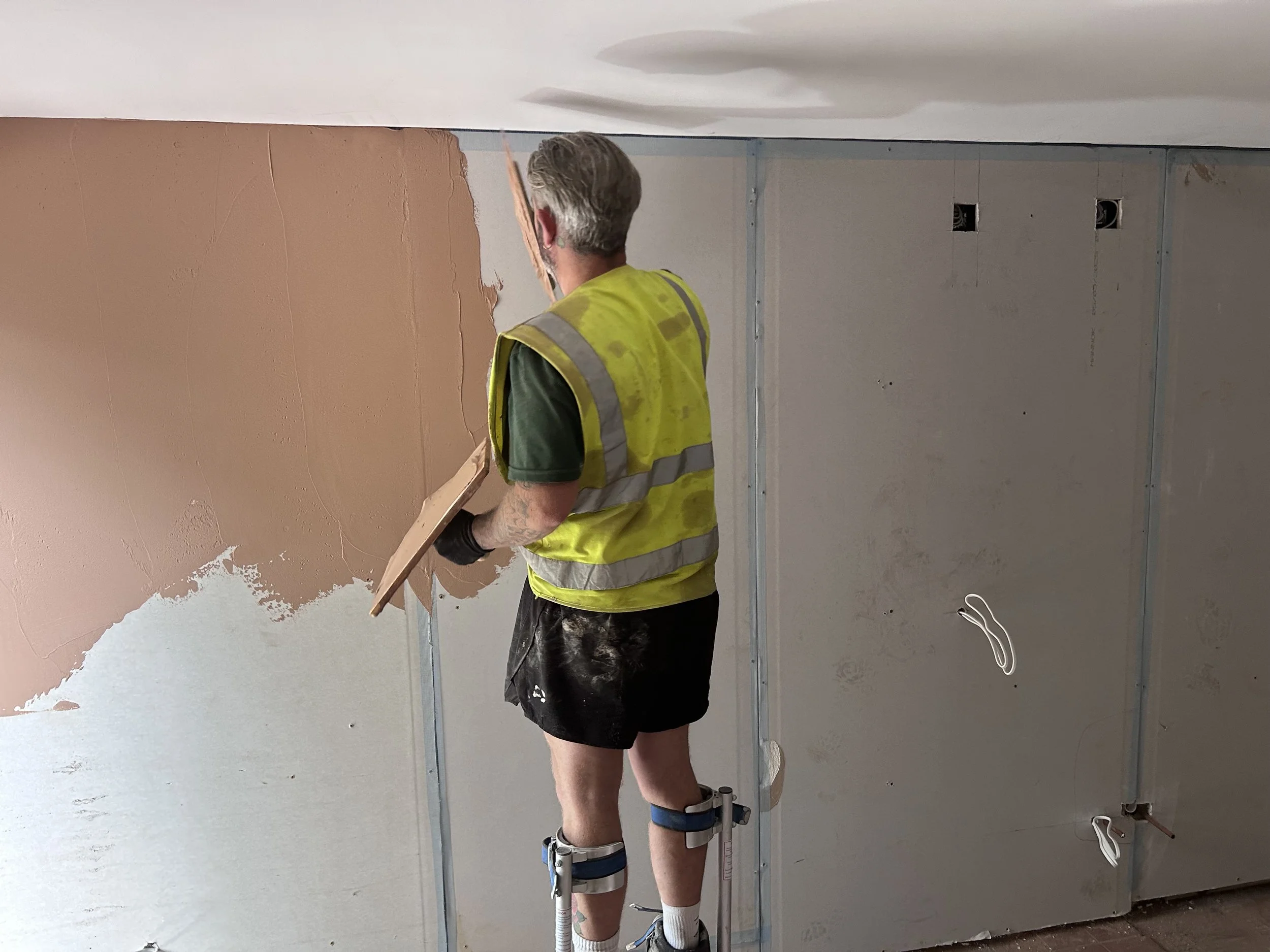 Construction worker in yellow safety vest and shorts installing drywall inside a room.
