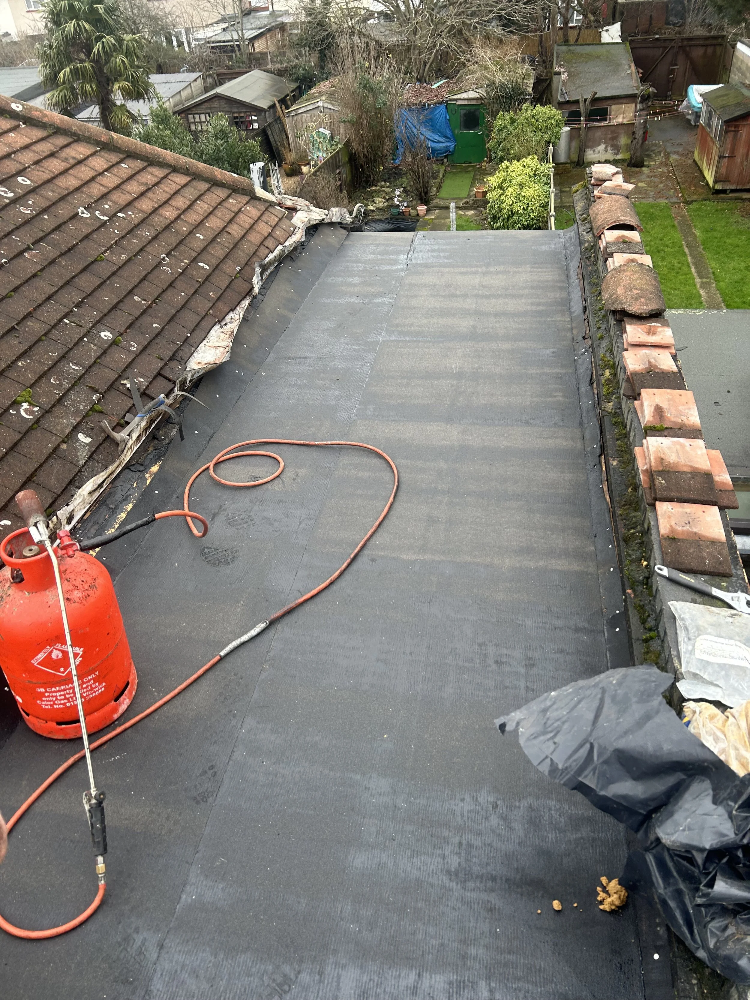 View from a rooftop showing a flat roof section being cleaned or coated, with a propane tank and hose, surrounded by older tiled roofs and a backyard with garden sheds, trees, and green lawns.