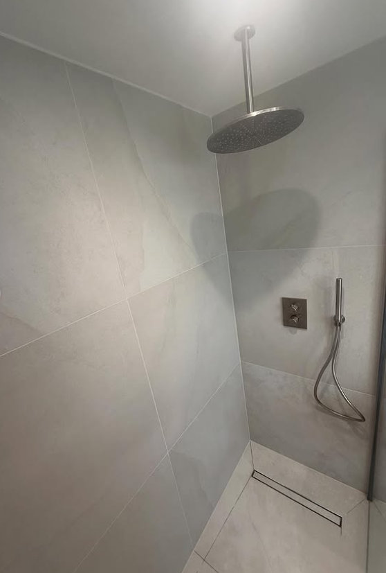 A modern shower with large light gray tiles on the walls and floor, a round overhead showerhead, a wall-mounted control panel, and a handheld showerhead with a hose.