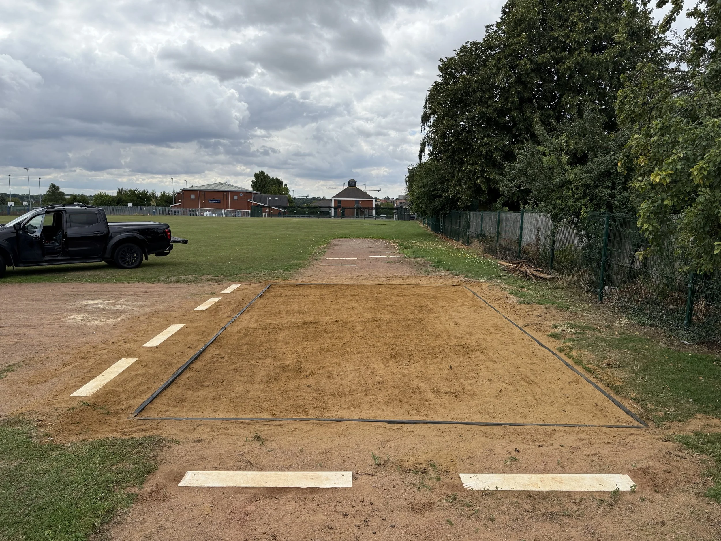 A sports field with a dirt track for shot put or discus, marked with black lines, surrounded by grass, with a black pickup truck parked on the grass to the left, and a large tree on the right. There are buildings and a cloudy sky in the background.