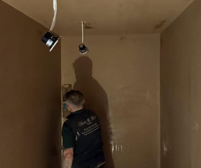 A person painting or preparing a wall in a room with two ceiling light fixtures hanging from the ceiling.