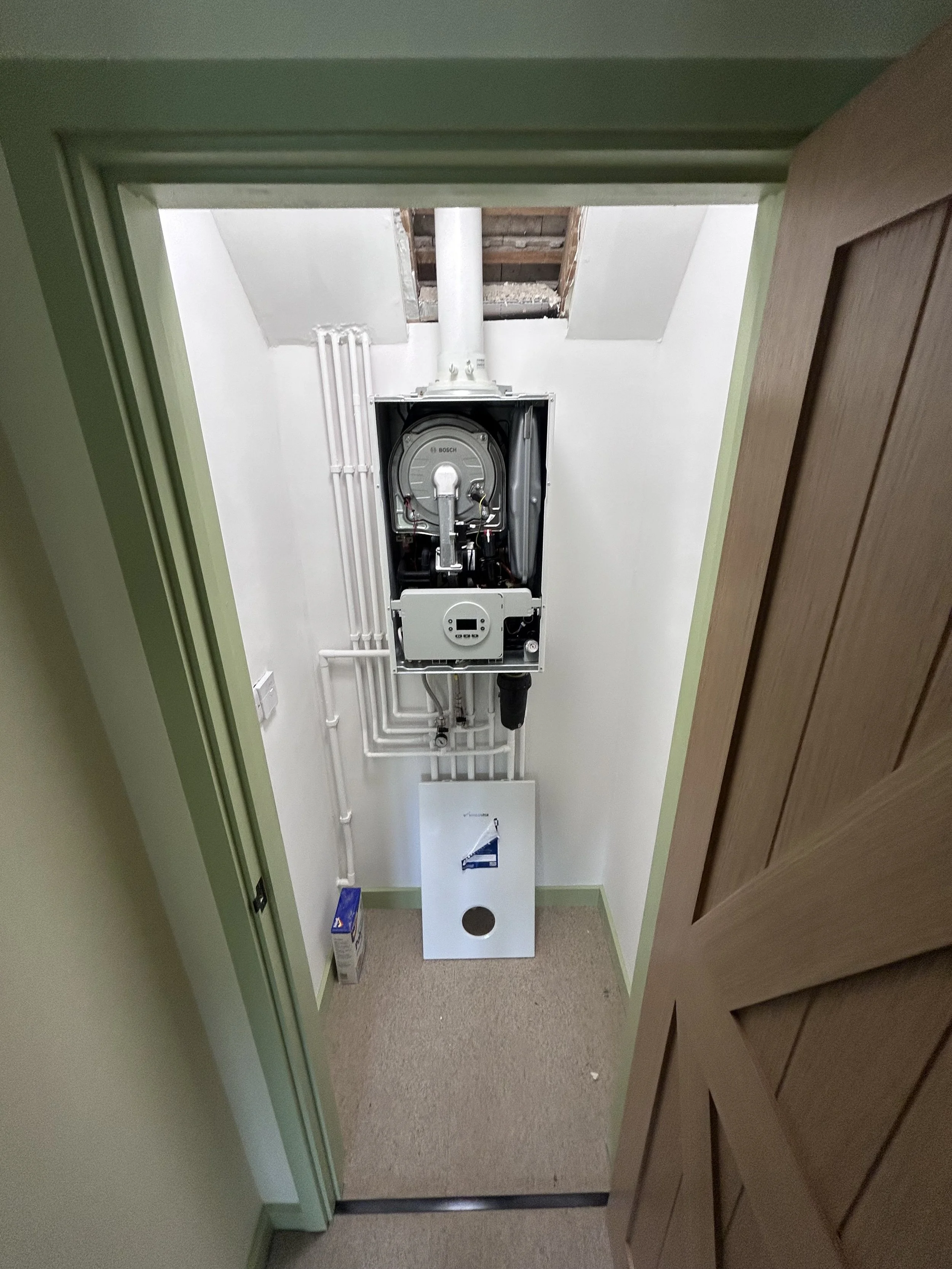 View of a laundry room with a wall-mounted boiler, pipes running along the wall, a cardboard box, and a wood-paneled door partially open.