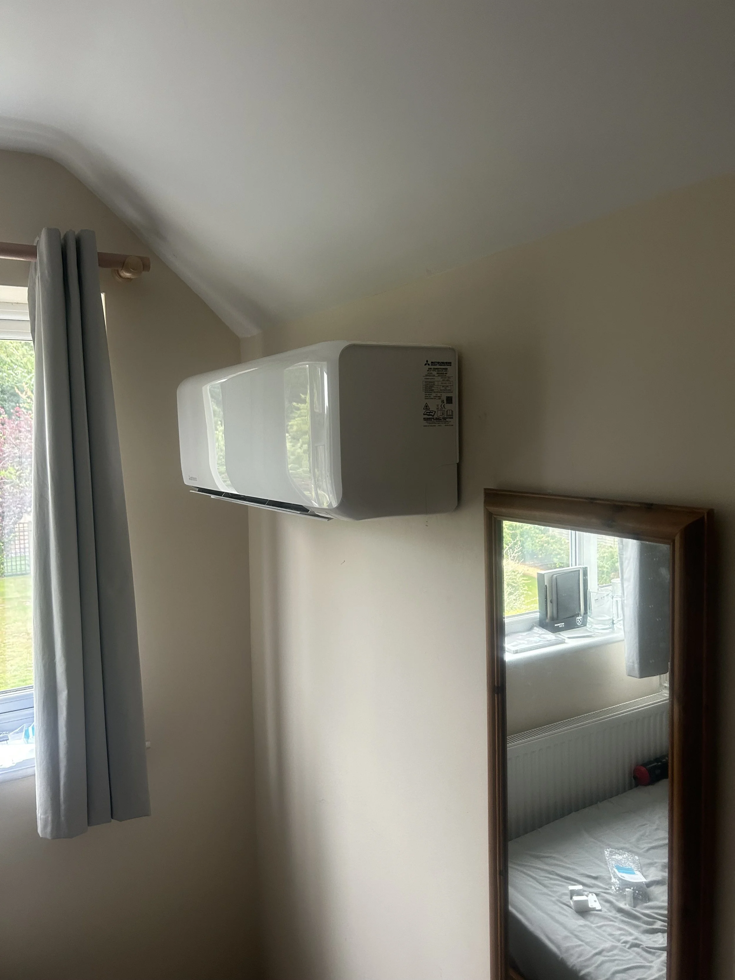 Wall-mounted air conditioner unit in a bedroom, with a window and curtain on the left and a mirror reflecting a bed and window on the right.
