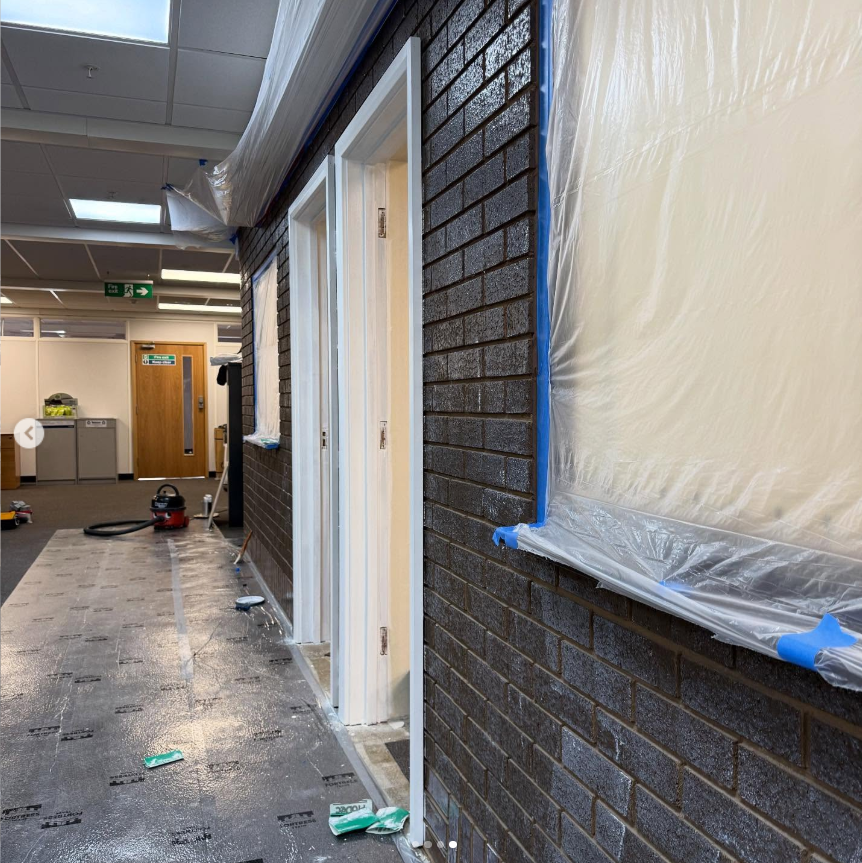 Interior wall undergoing renovation with brickwork, window, and door frames, covered in plastic sheeting, on floor are tools and debris.