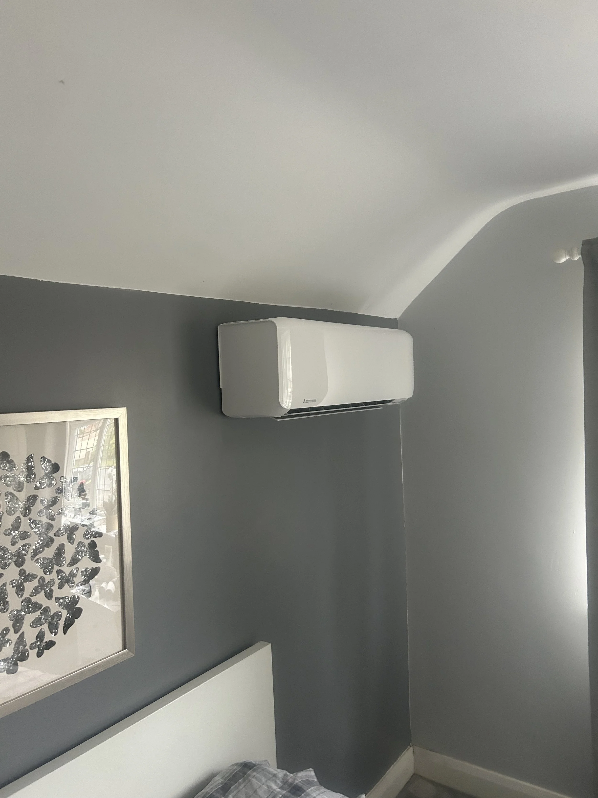 Indoor bedroom corner with dark gray walls, a white air conditioning unit mounted on the wall, a framed butterfly artwork, part of a bed with a plaid blanket, and a curtain on the window.