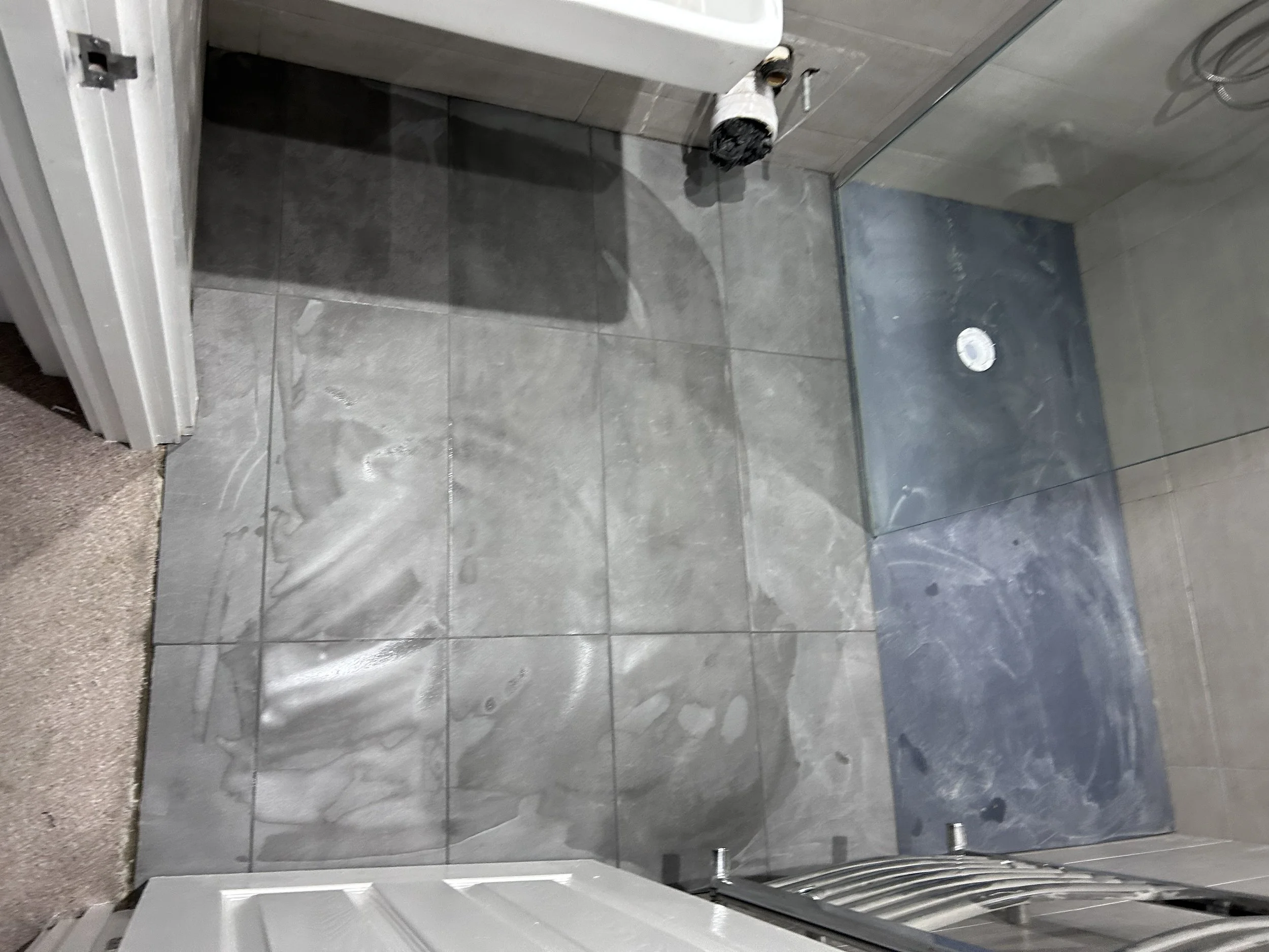 Bathroom with gray tiles and a partially installed shower area, including a metal drain and a plastic wrapping on fixtures.