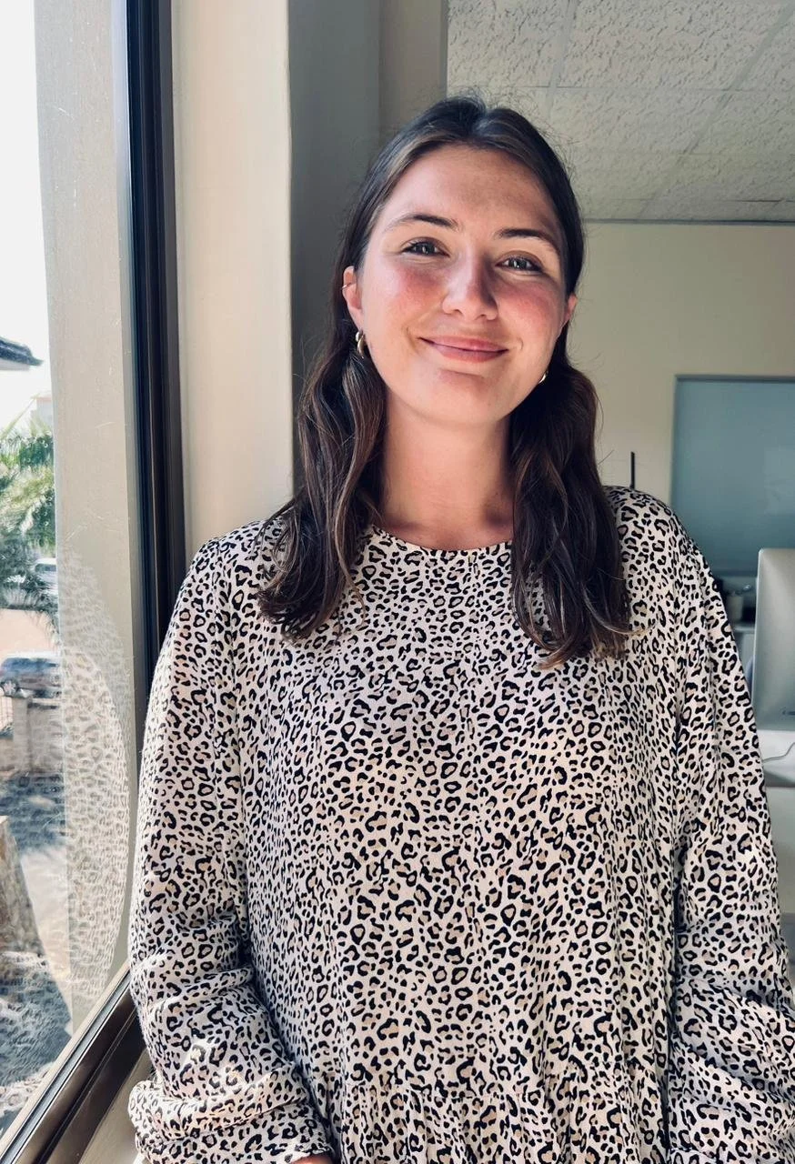 A woman with shoulder-length brown hair, wearing gold hoop earrings and a leopard print top, standing near a window indoors with a smile.
