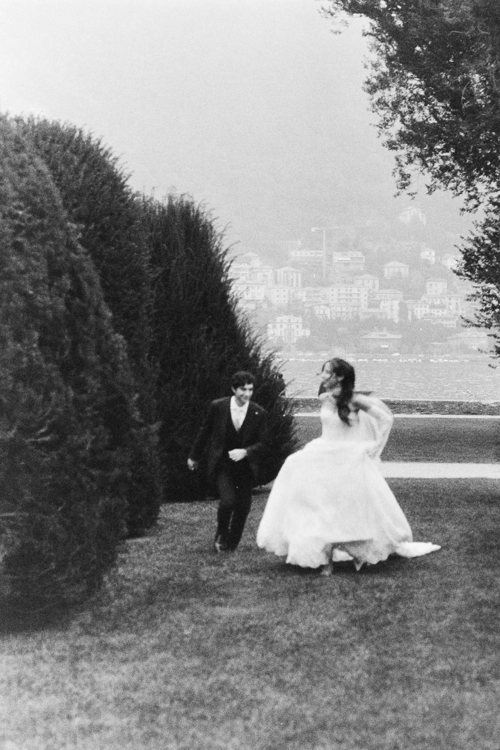 Grainy black and white 35mm film photo of a bride and groom running and laughing on the grass at Lake Como.