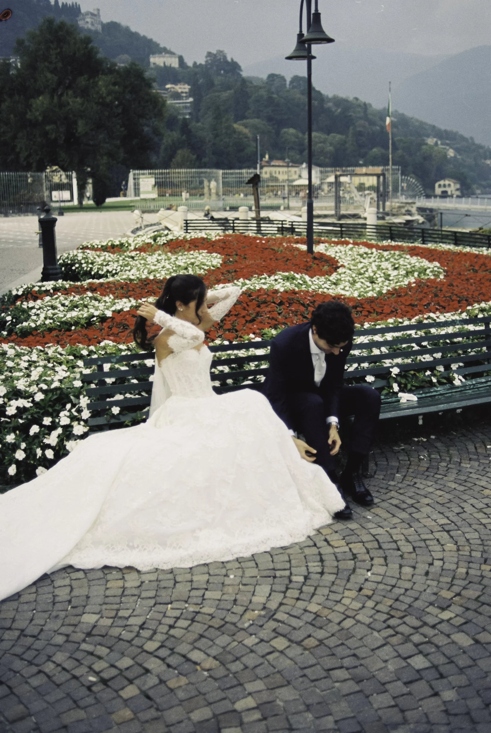 Candid 35mm film photo of a bride and groom sitting on a park bench at Lake Como, groom tying his shoe.