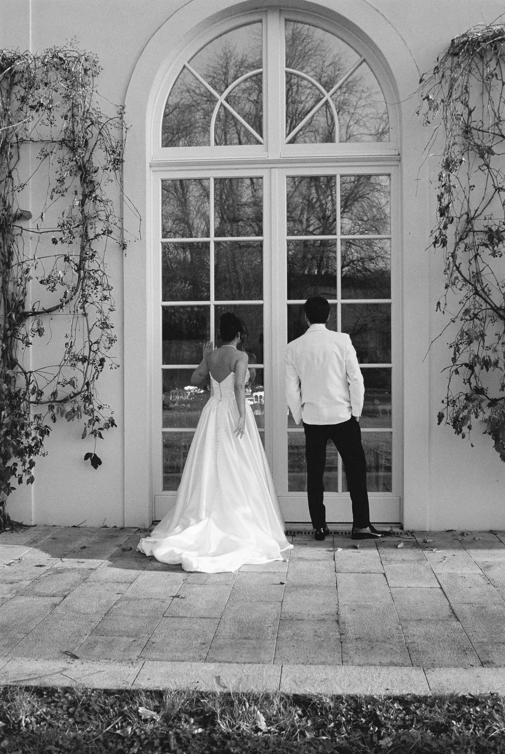 Black and white 35mm portrait of bride and groom from behind, looking out a large arched glass portal at Schloss Lübbenau.