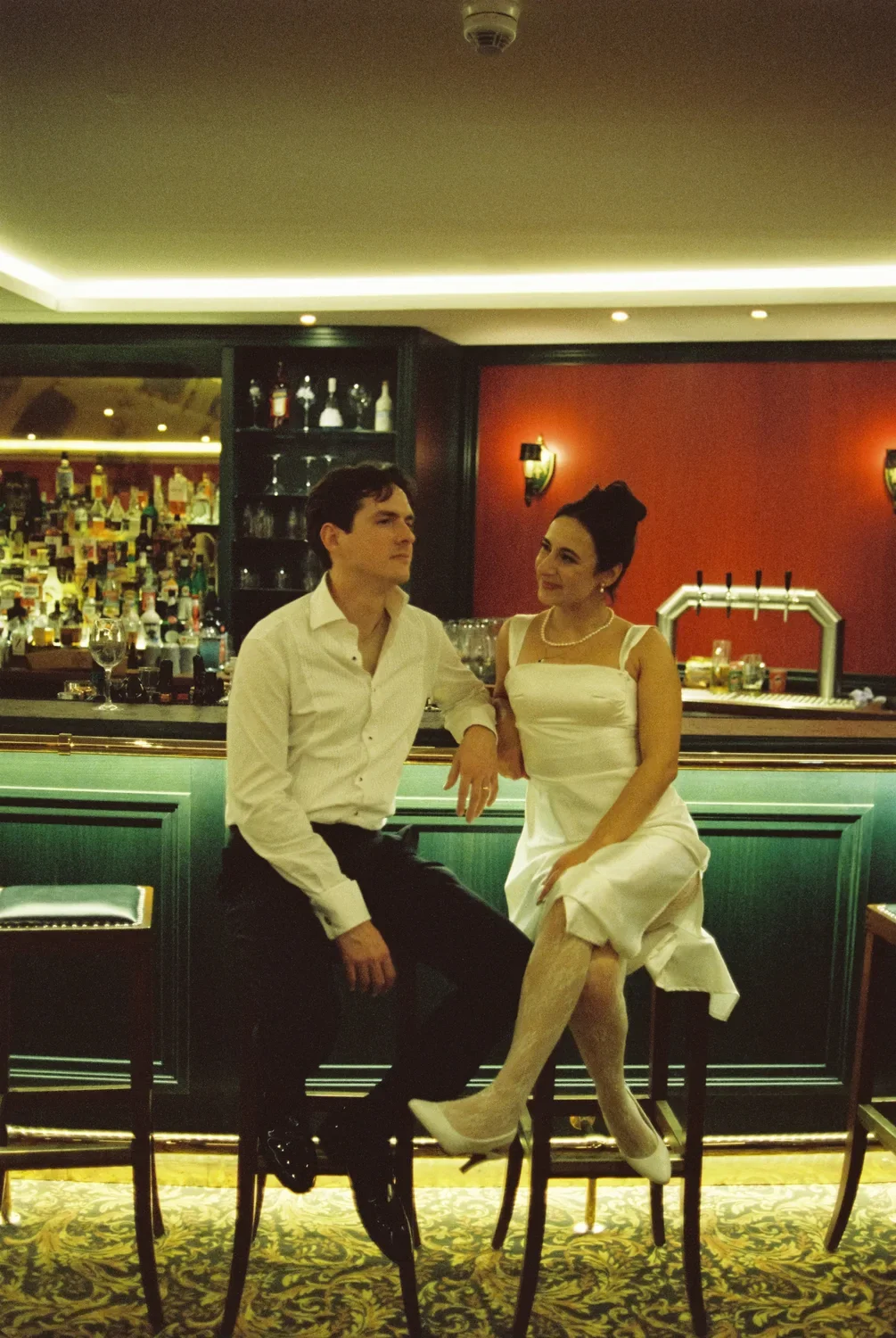 A quiet moment at the bar: Munira and Felix sitting on stools, looking at each other away from the party.