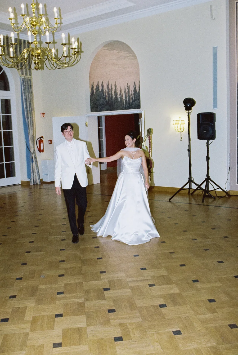 Wide shot of the bride and groom holding hands as they enter a vast ballroom with a crystal chandelier.