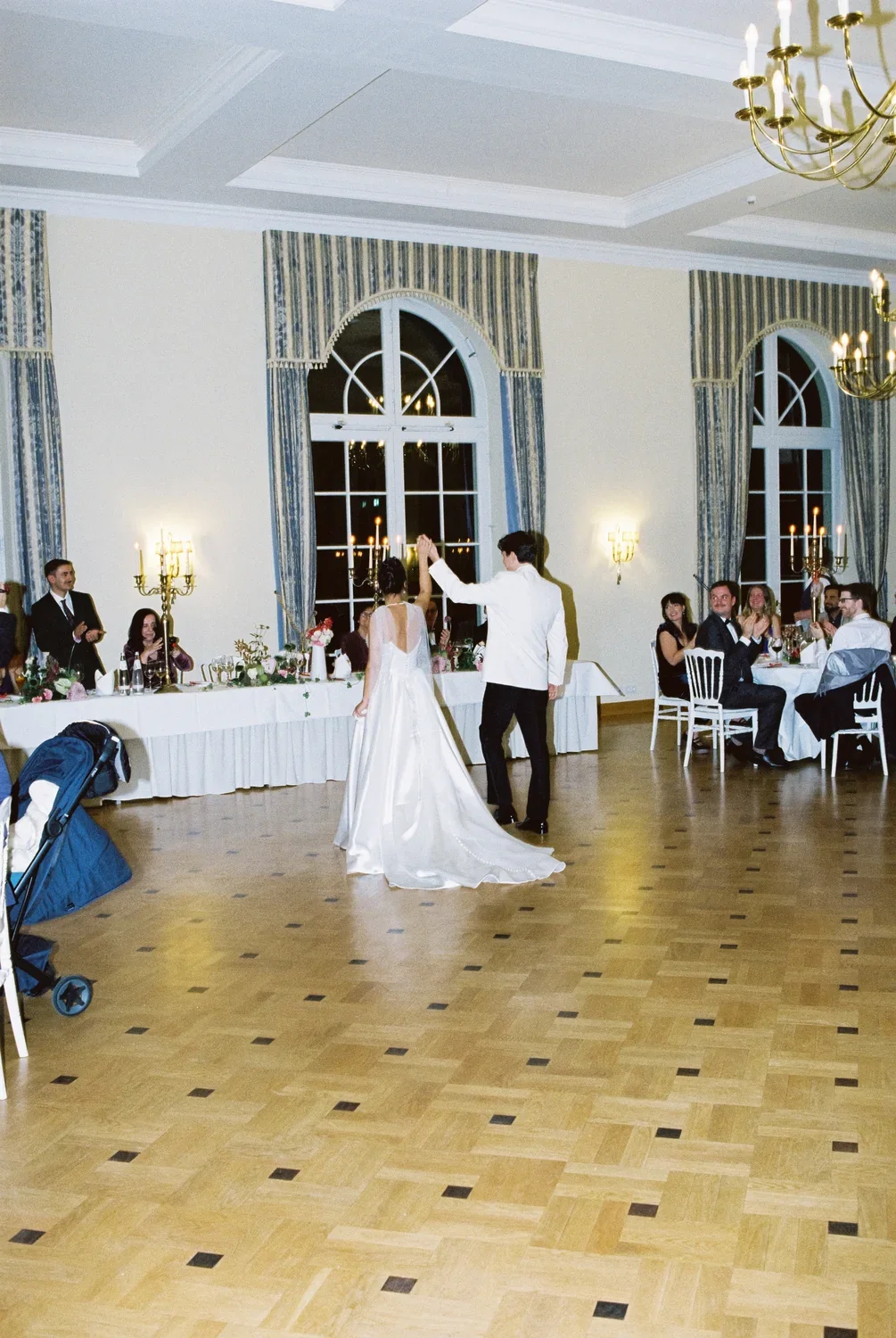 Rear view of the couple cheering with raised hands in the middle of a ballroom, guests applauding at their tables.