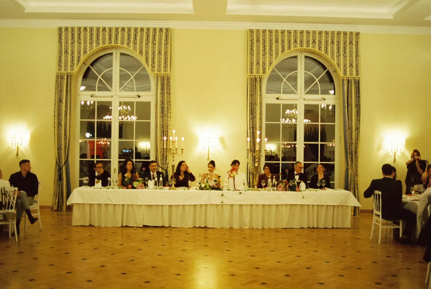 A symmetrical shot of the wedding party at a long white-clothed head table beneath arched windows.